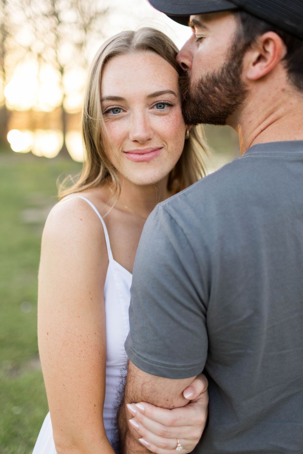 A bearded man in a gray t-shirt kisses a smiling blonde woman with freckles outdoors at golden hour.