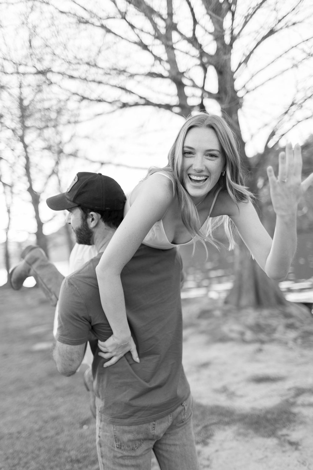 Black and white photo of a laughing woman getting a piggyback ride from a bearded man in a cap outdoors.