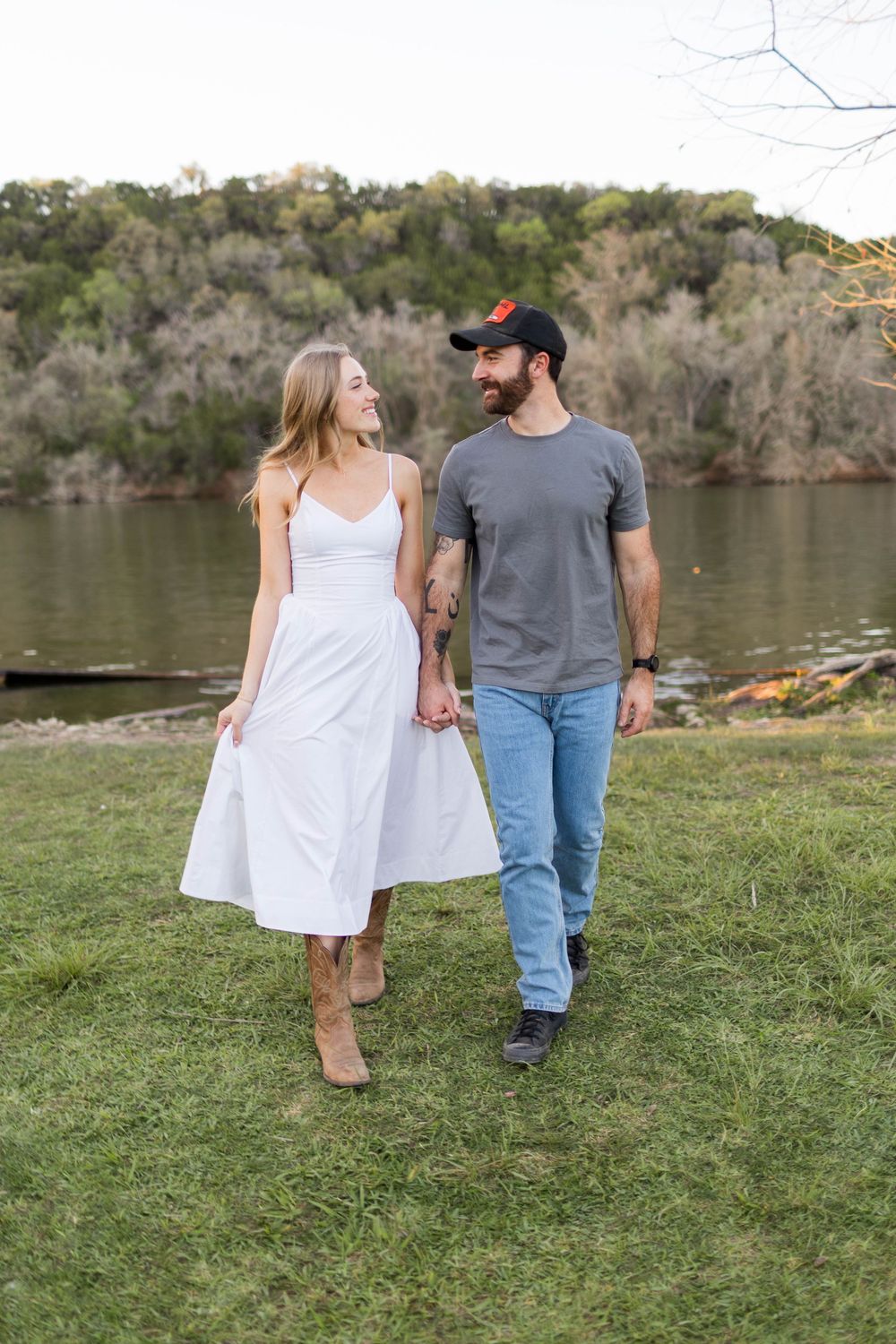 A couple holds hands while walking on grass near a river, woman in white dress and man in gray tee and jeans.