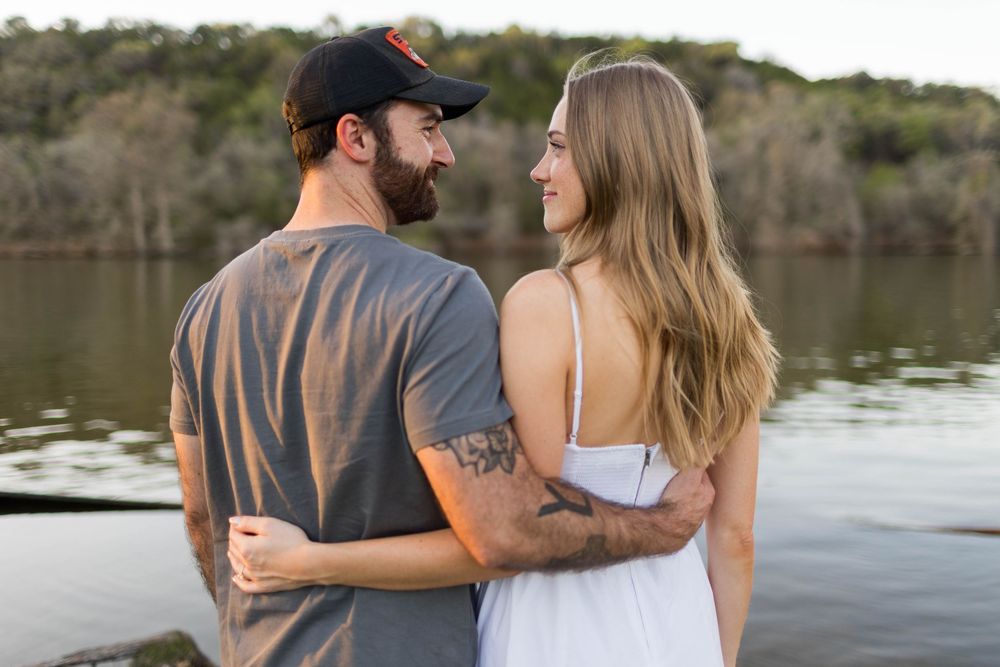 A couple embraces by a lakeside, the man in a gray tee and cap, the woman in a white dress, sharing a romantic moment.