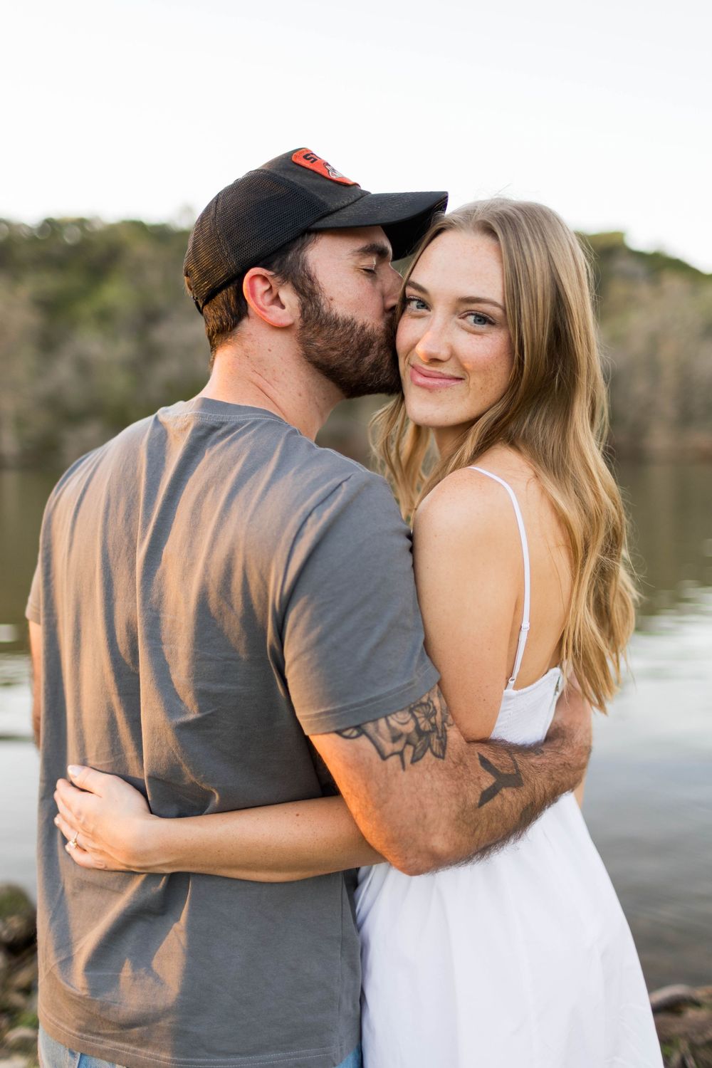 A couple shares a tender kiss by a lake, man in gray tee and cap, woman in white dress with tattoos.
