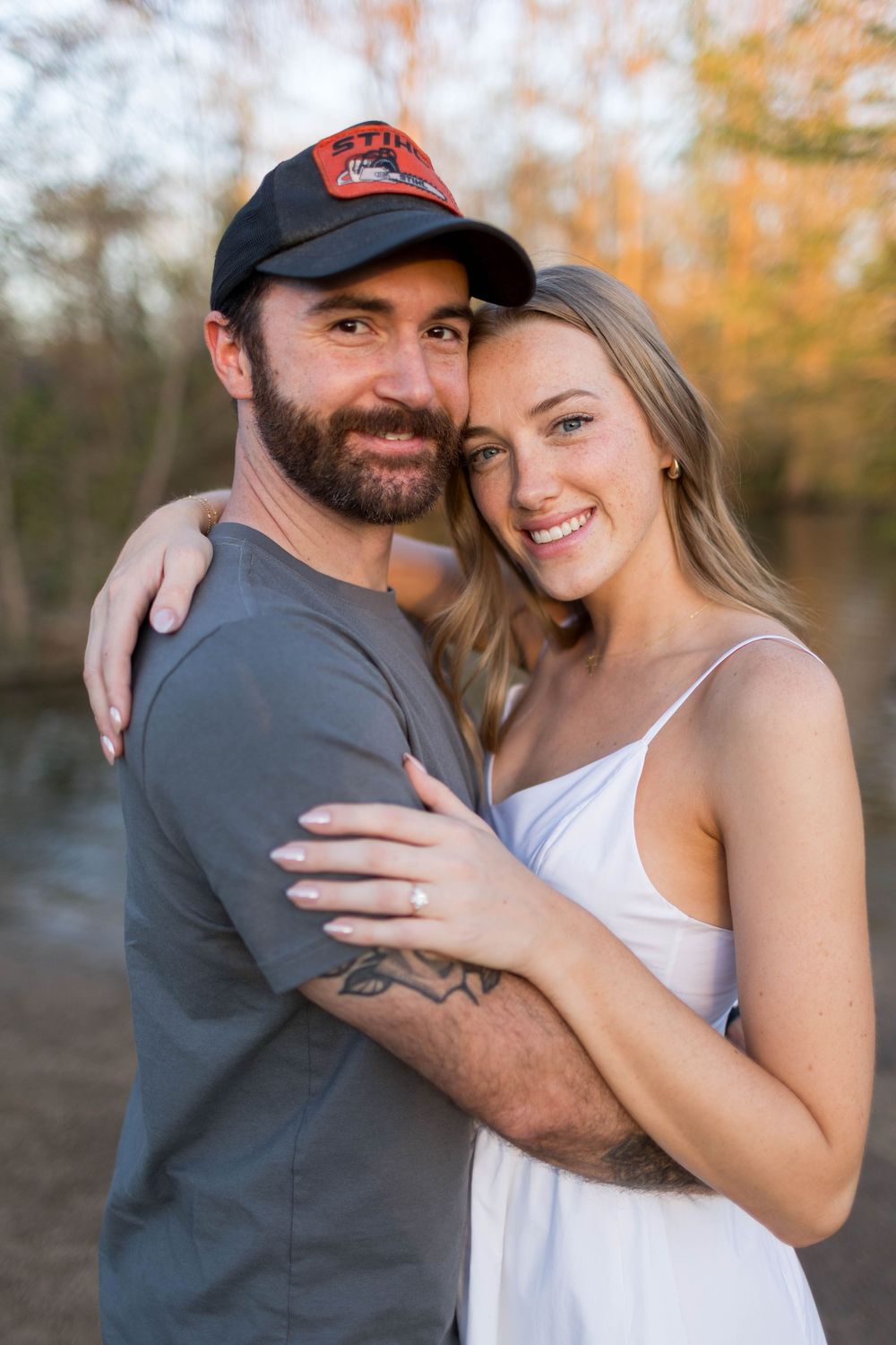 A smiling couple embraces outdoors, man in gray tee and black cap, woman in white dress, autumn trees in background.