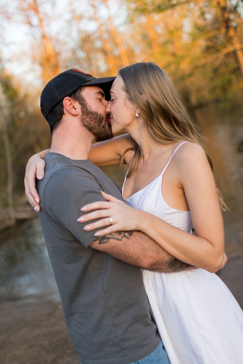 A couple sharing a romantic kiss outdoors near a creek, surrounded by autumn trees in warm golden light.