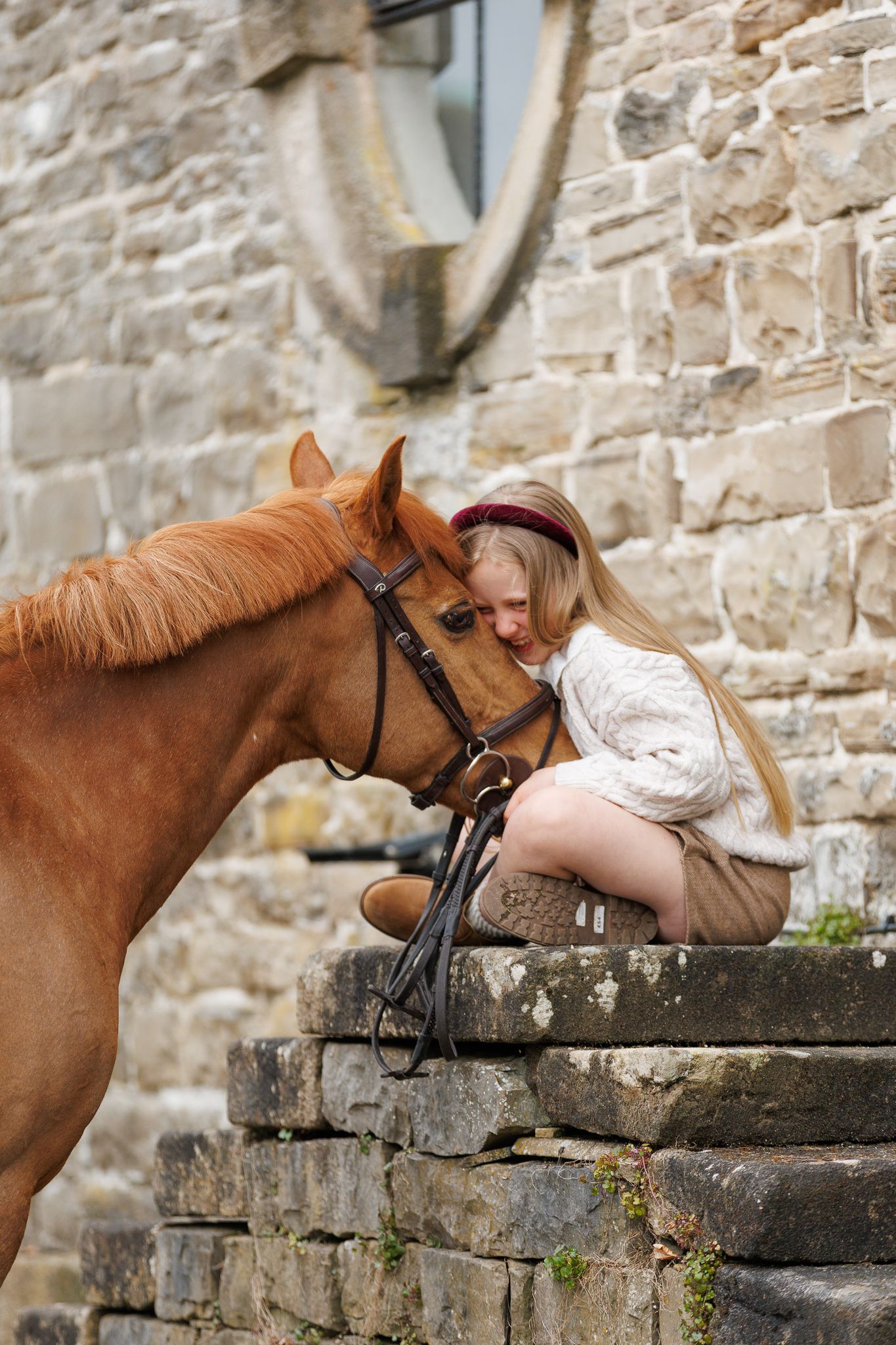 A young blonde girl sits on stone steps, tenderly leaning her head against a chestnut horse wearing a bridle.