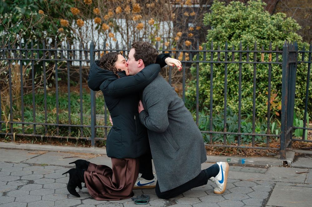 Two people sharing a romantic kiss on a city sidewalk near an iron fence surrounded by autumn foliage.