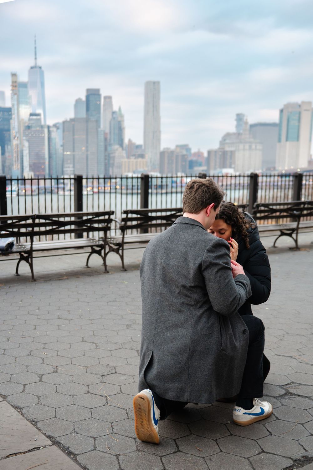 Man on one knee proposing to woman at NYC waterfront with Manhattan skyline backdrop.