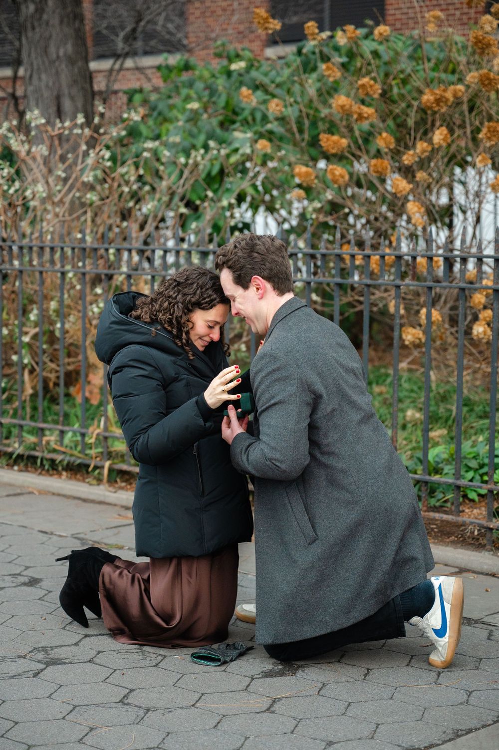 Man kneeling on one knee proposing to woman in winter coats on a cobblestone path in an urban park.