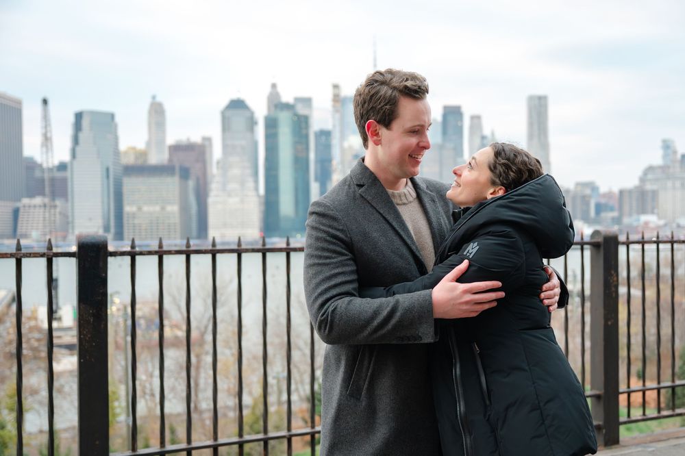 Couple embracing and smiling at each other with New York City skyline backdrop on a cloudy day.