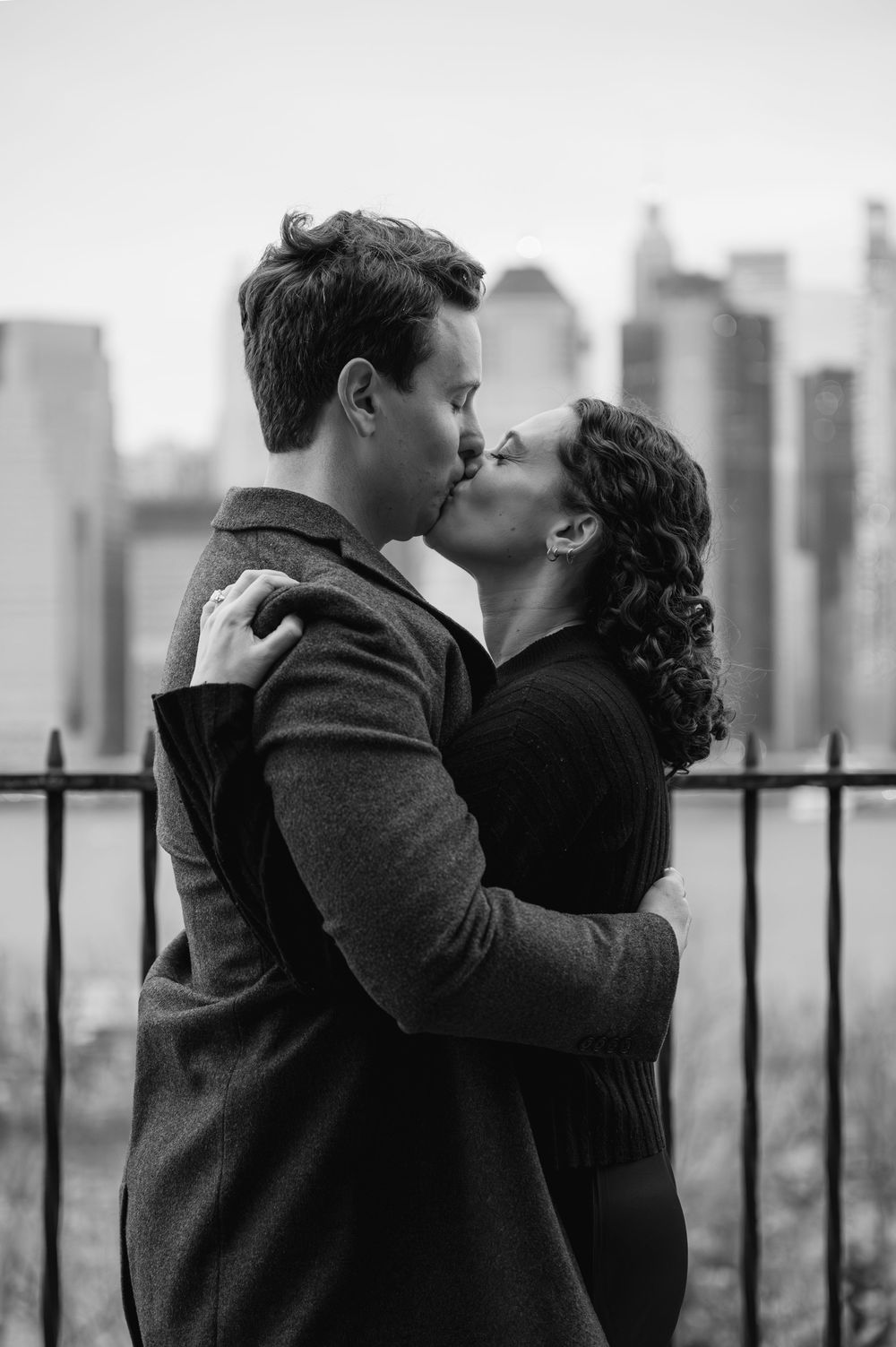 Black and white photo of a couple kissing with a city skyline backdrop, embracing romantically outdoors.