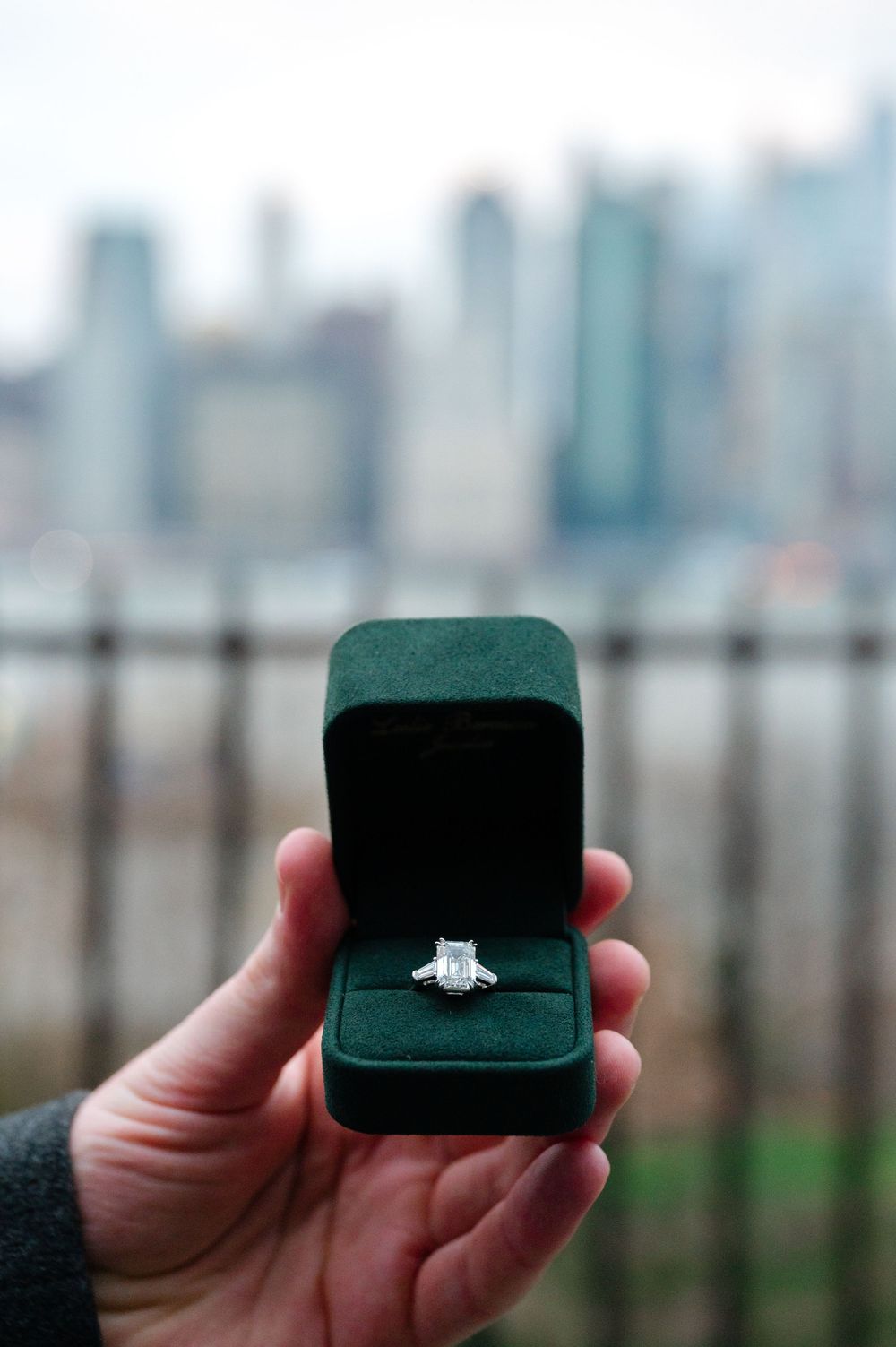 Hand holding a green velvet box with a diamond engagement ring, NYC skyline in the background.