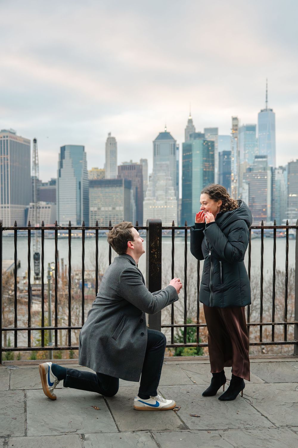Man proposes on one knee to woman with NYC skyline backdrop at Brooklyn Bridge Park.