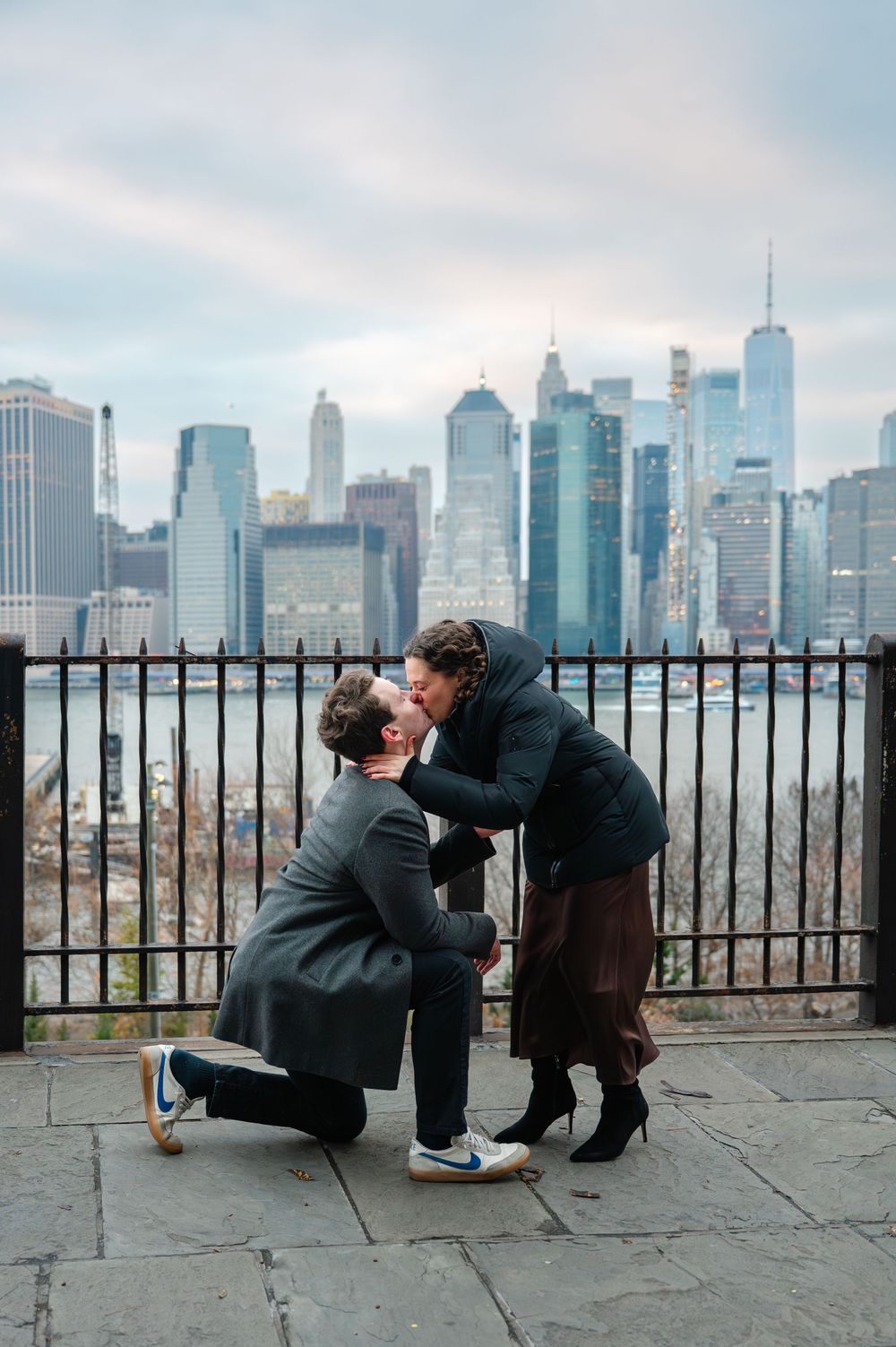 A couple shares a kiss during a marriage proposal on one knee with the NYC skyline backdrop.