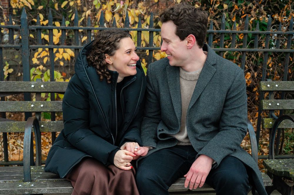 A smiling couple sits on a park bench holding hands, surrounded by autumn foliage and iron fence.