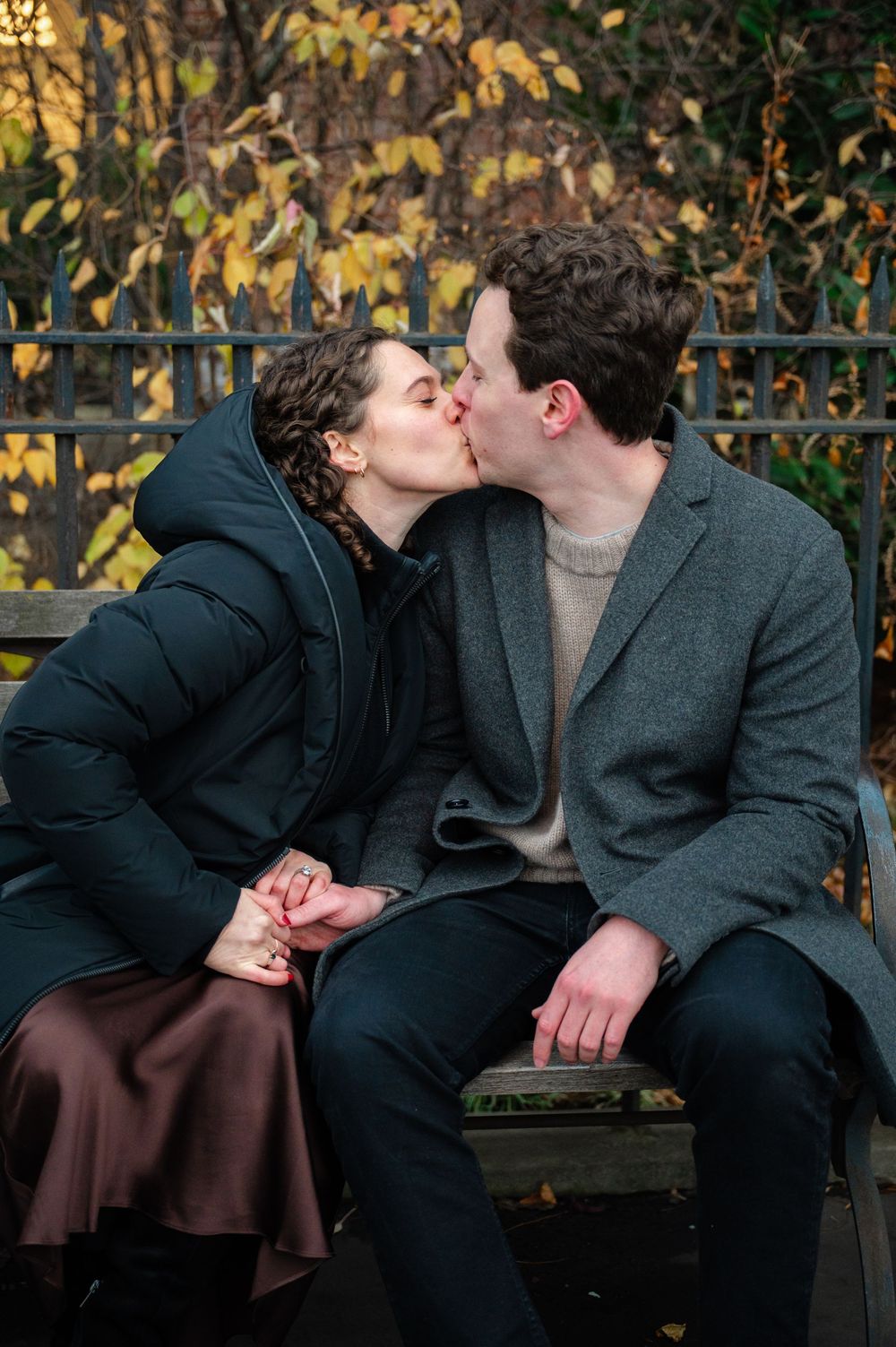 Couple kissing on a bench in autumn park, woman in puffer coat, man in grey wool coat.