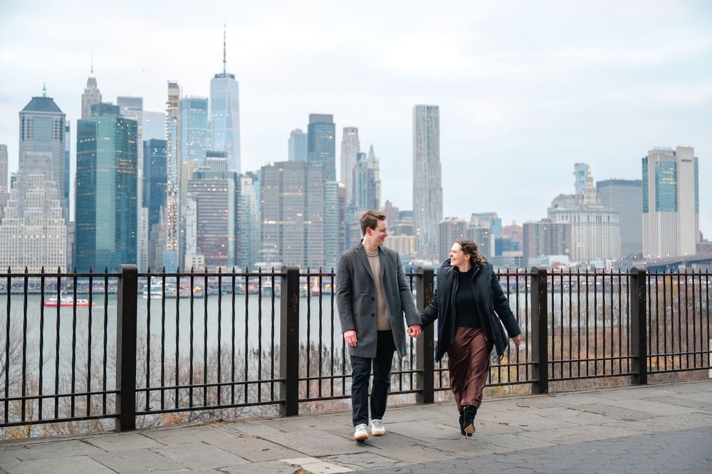 Couple walking hand in hand along a waterfront promenade with the NYC skyline behind them.