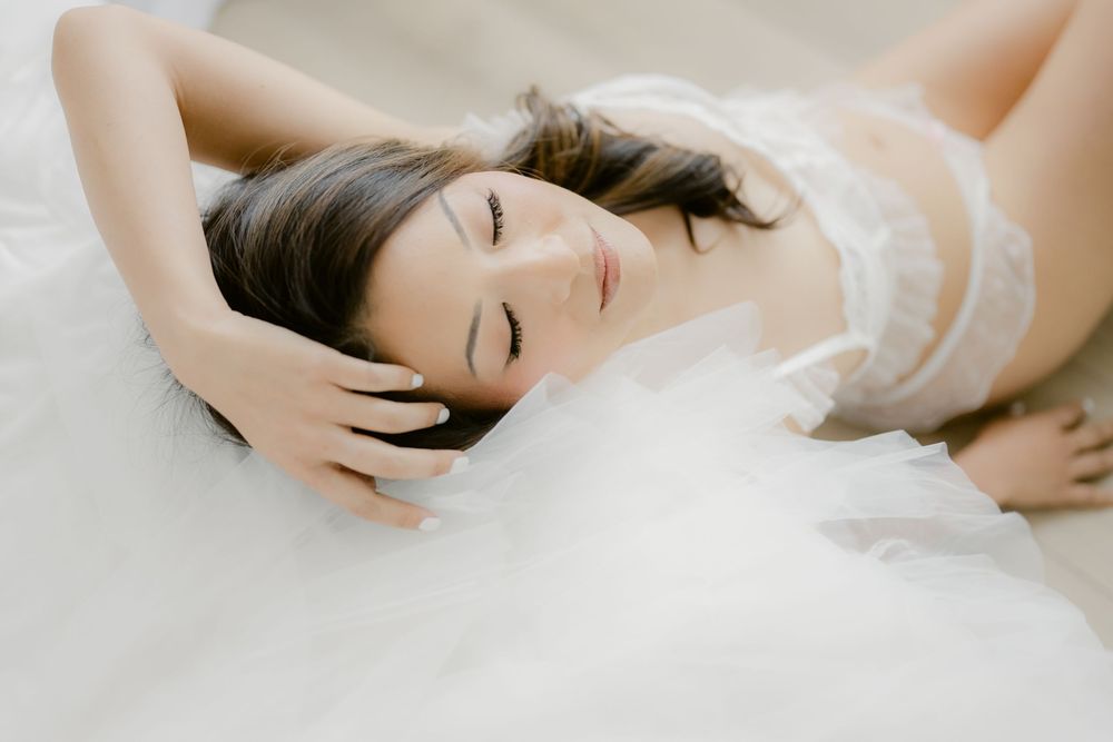 Woman in white lace lingerie lying gracefully on a soft white tulle fabric with eyes closed.