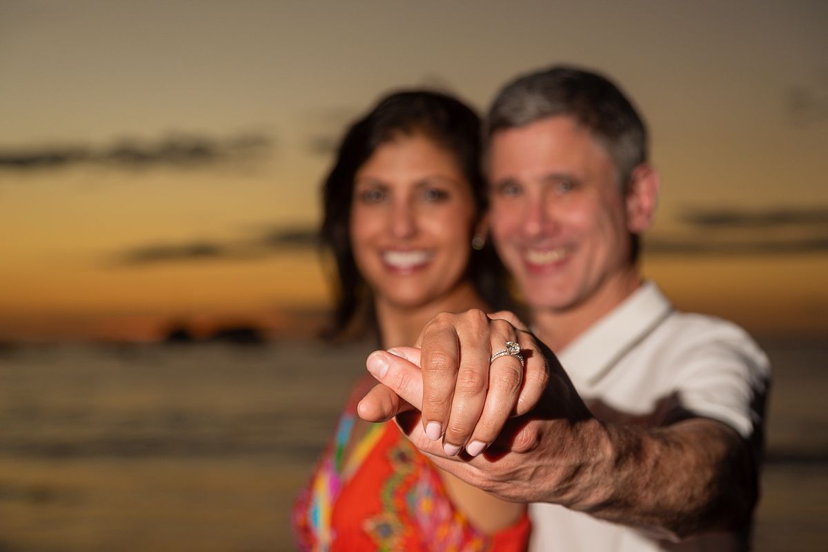 Couple holding hands showing engagement ring at sunset beach, romantic proposal moment with golden sky backdrop.