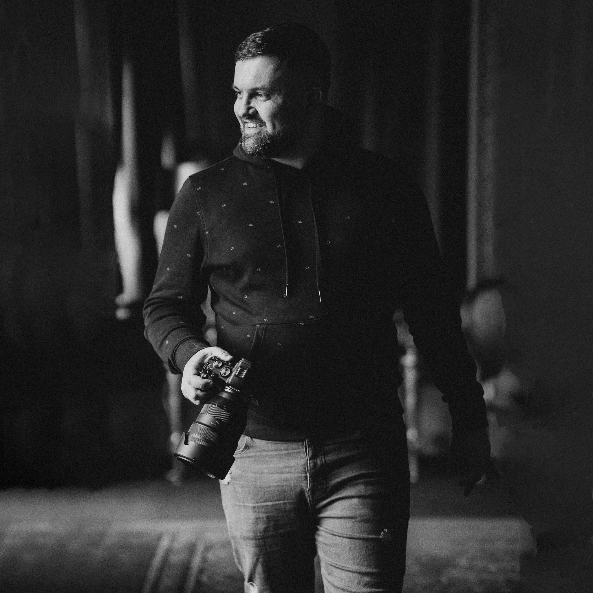 Black and white photo of a bearded man holding a camera, smiling in a dimly lit indoor setting.