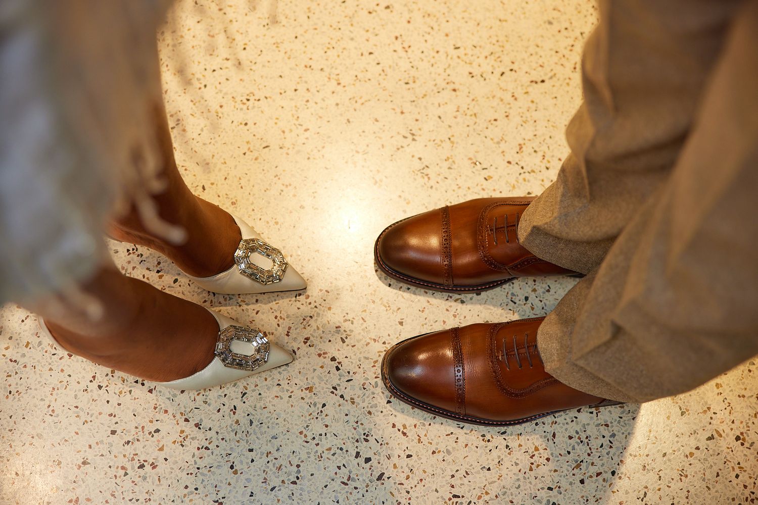 Top-down view of a bride and groom's feet showing brown leather dress shoes and silver bridal heels on a speckled floor.