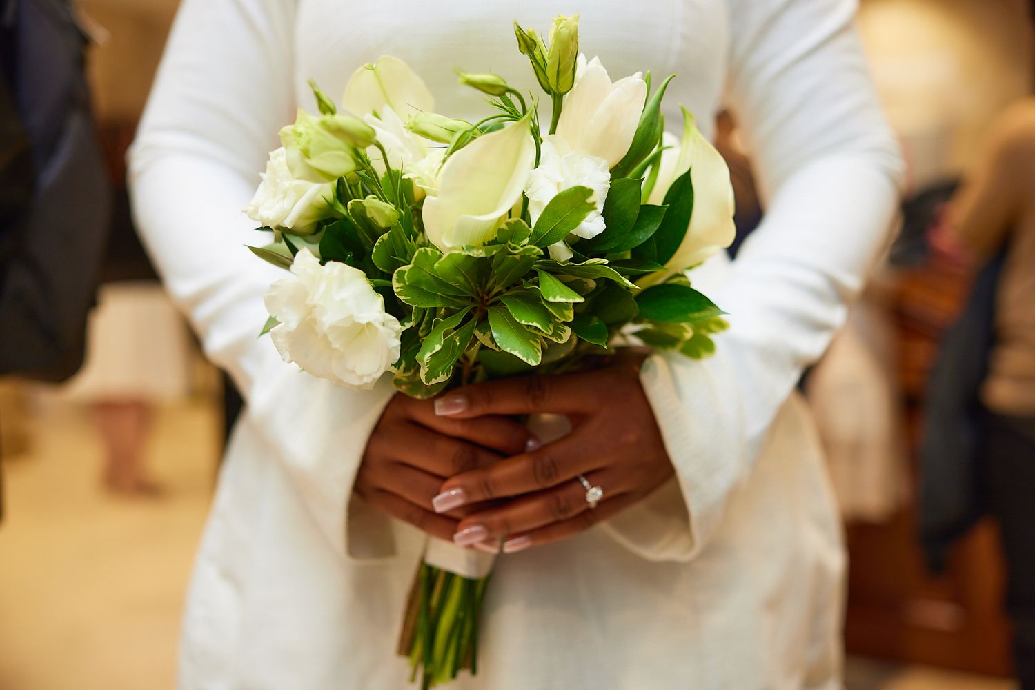 Bride in white dress holding elegant bouquet of white roses and green flowers, wearing wedding ring.