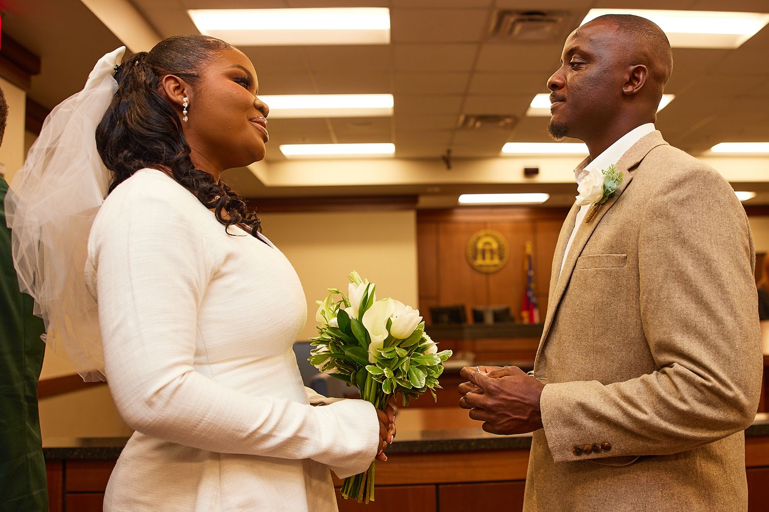 A couple exchanges vows in a courthouse wedding, bride in white gown with veil holding flowers, groom in tan suit.