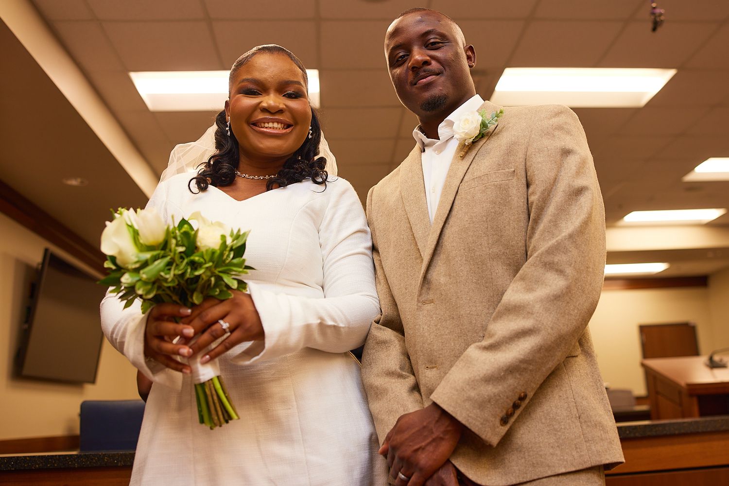 A smiling couple poses together on their wedding day, bride holding white bouquet in white dress, groom in tan suit.