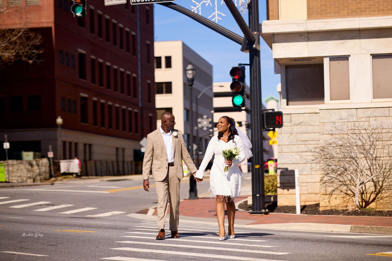 A Black bride and groom cross a city street holding hands, bride in white mini dress with bouquet, groom in tan suit.