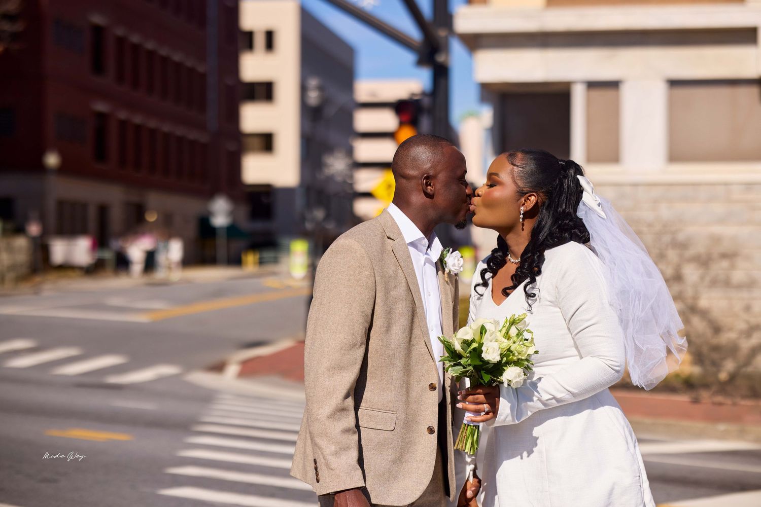 A newlywed couple shares a kiss on a city street, bride in white gown with veil holding bouquet, groom in tan suit.