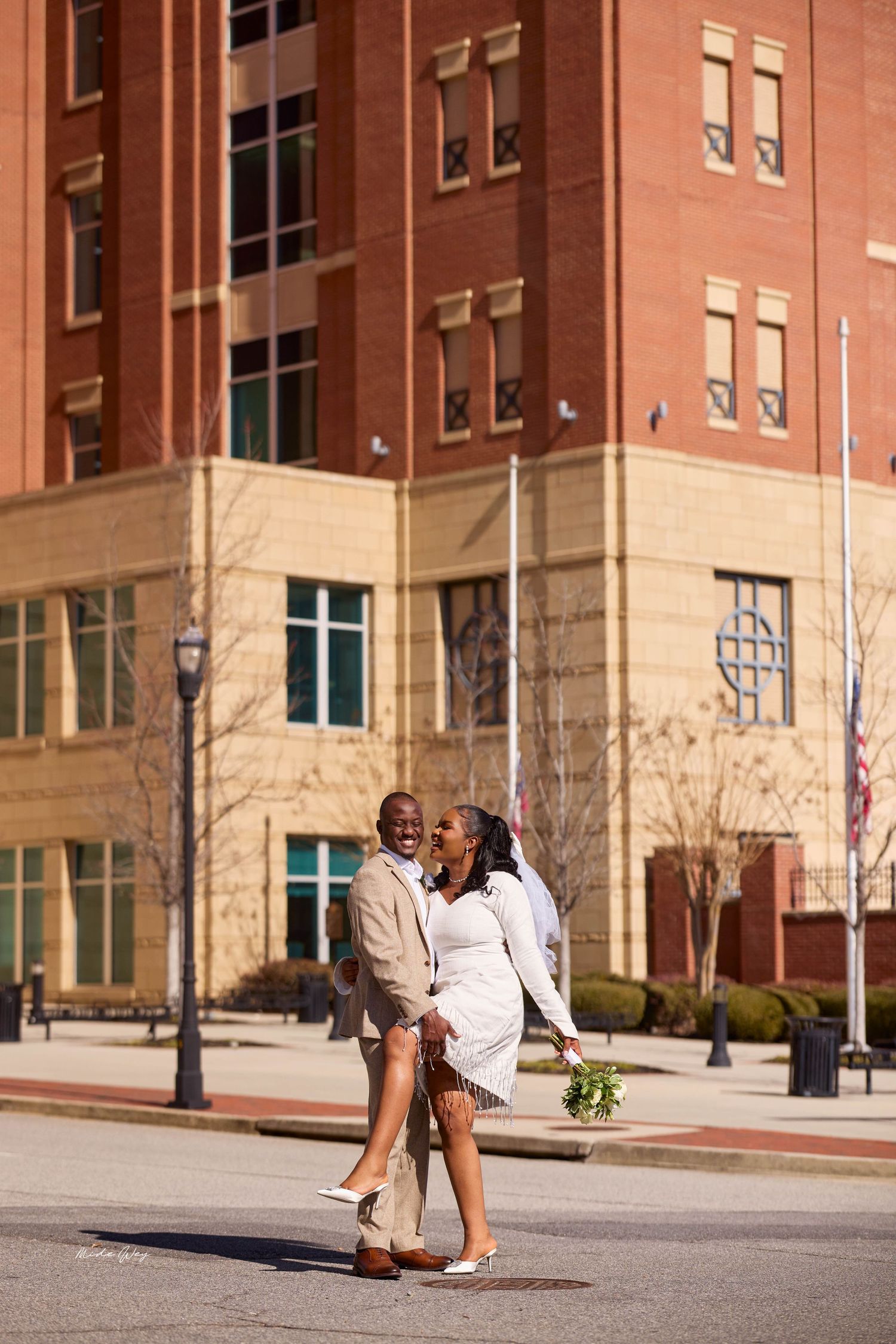 A joyful Black couple in wedding attire laughs together outside a red brick building on their wedding day.