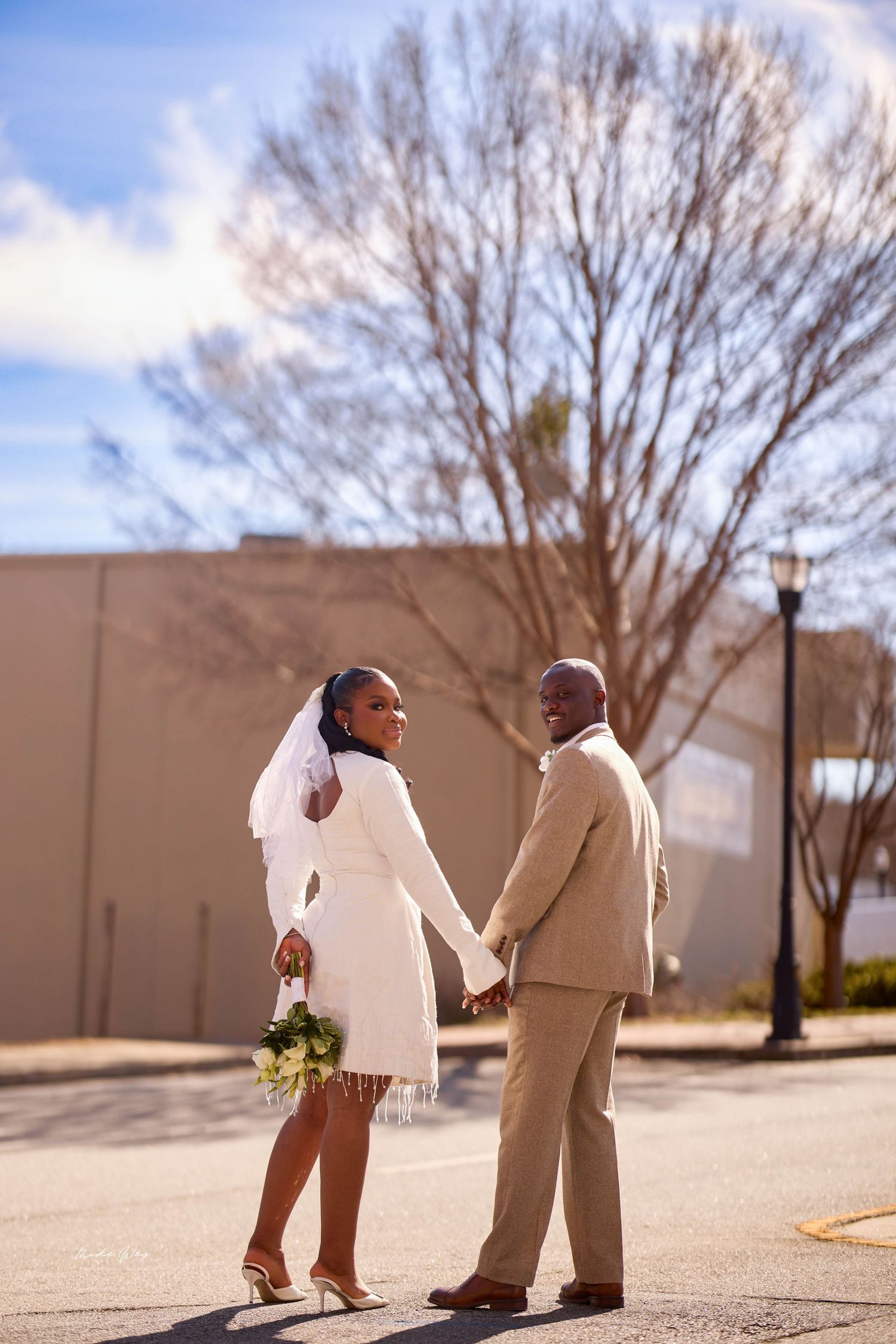 A Black bride in a white mini dress and veil holds hands with her groom in a tan suit outdoors.