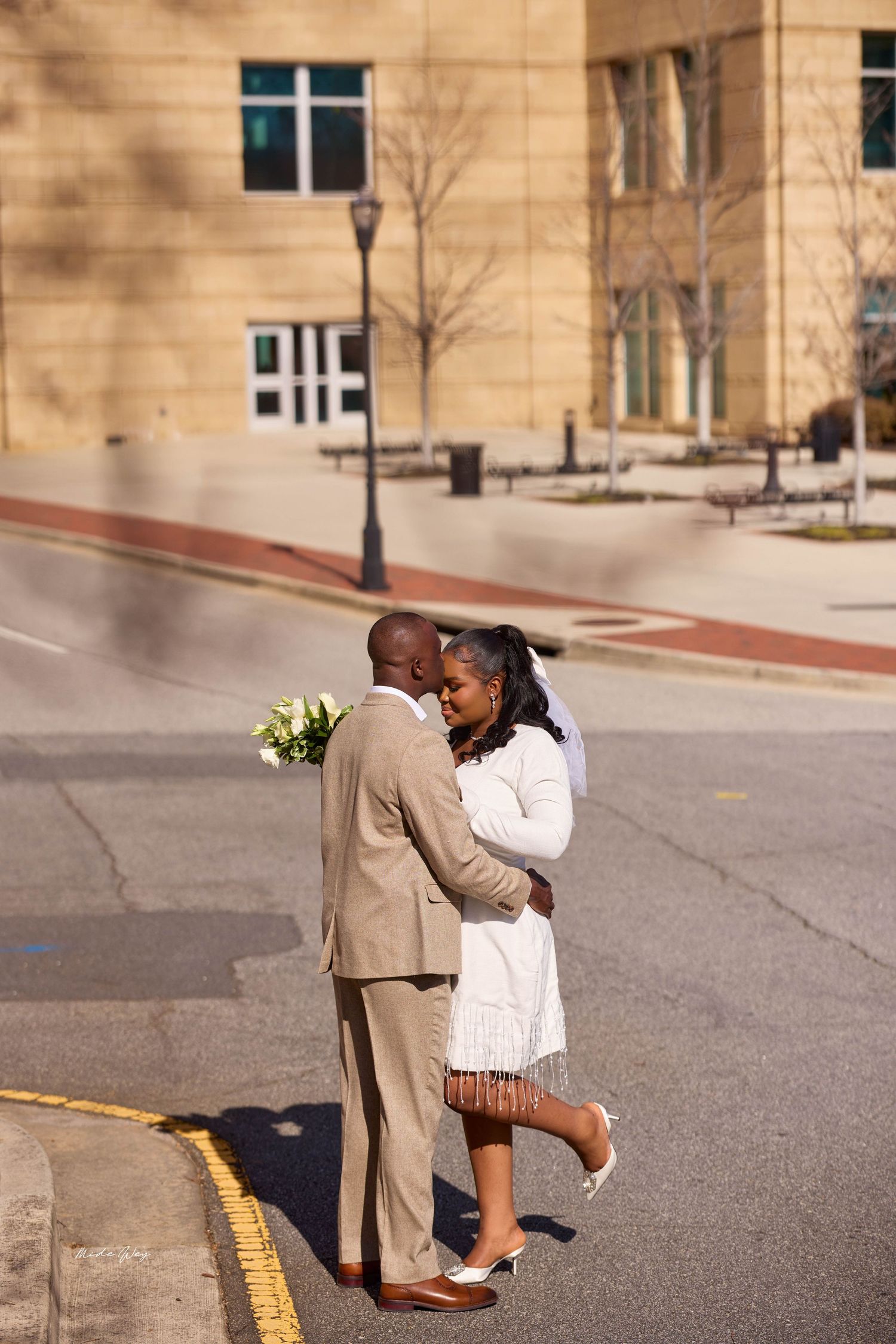 A newlywed couple embraces on a sunny street, groom in tan suit holding white bouquet, bride in white dress.