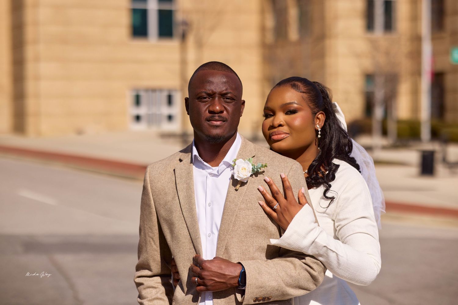 A couple in wedding attire poses outdoors, the groom in a tan suit with boutonniere and bride in white jacket.