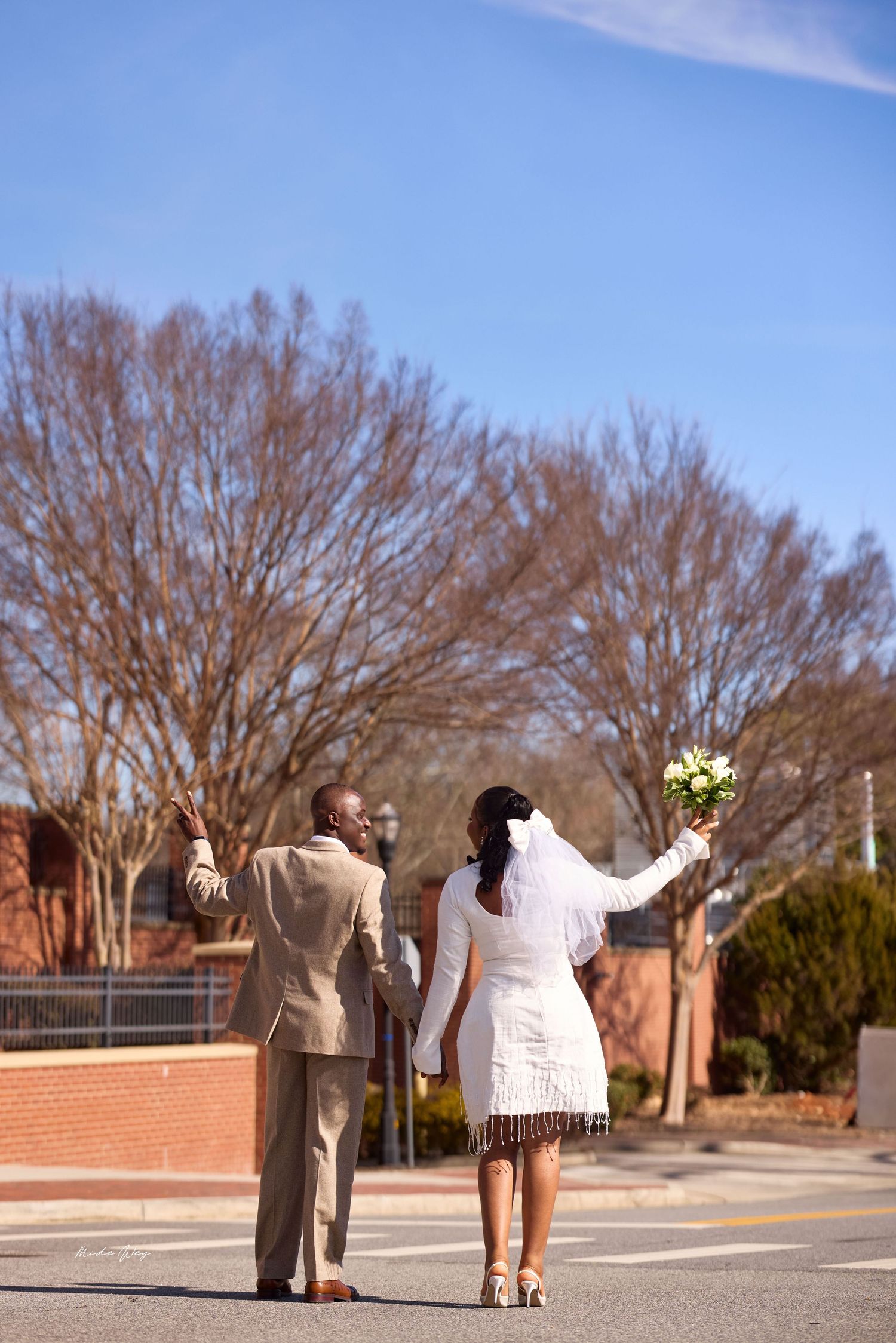Newlywed couple walking down street, bride in white dress holding bouquet, groom in tan suit, both raising hands.