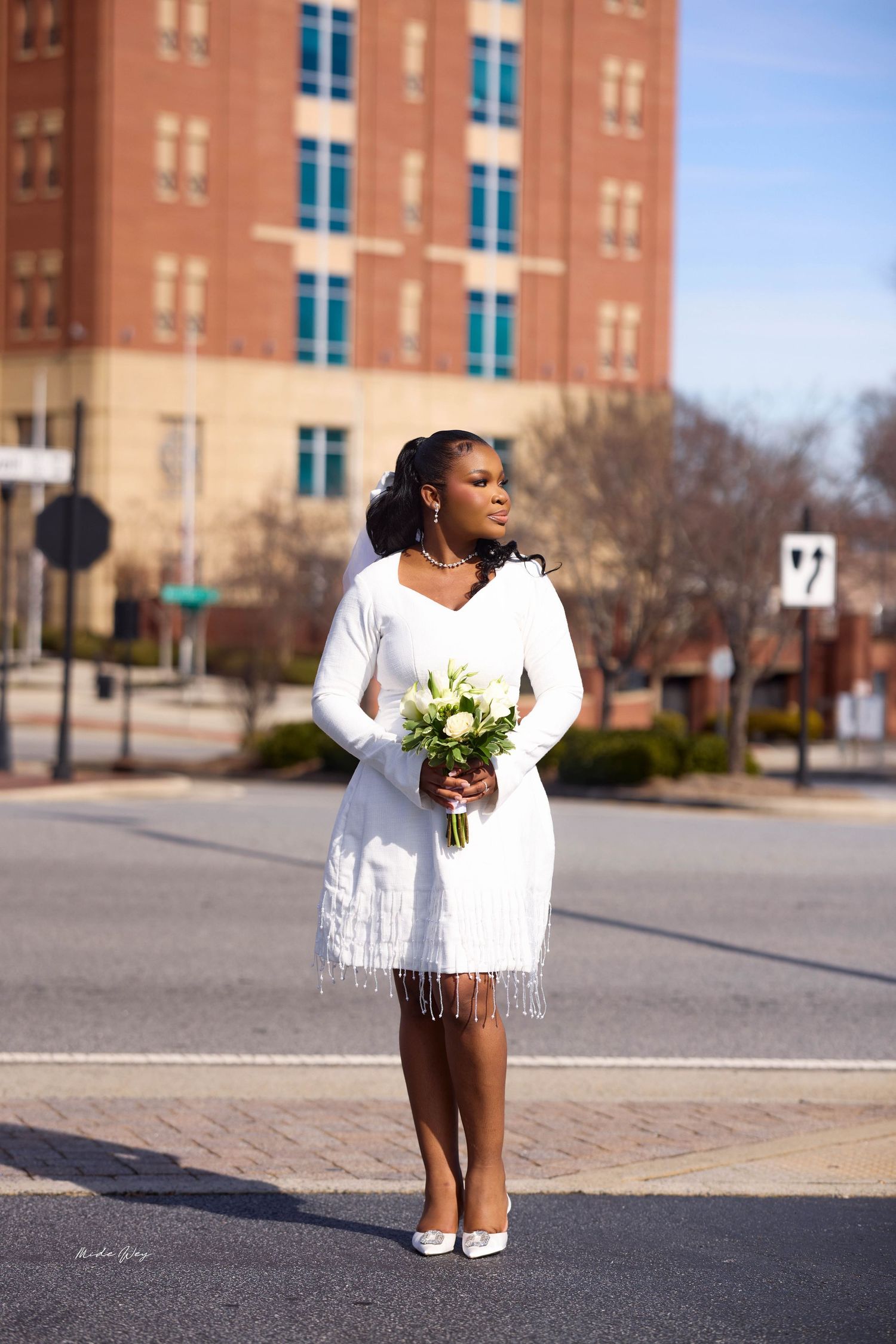 Woman in white fringe dress holding bouquet stands on urban street corner near brick building on sunny day.