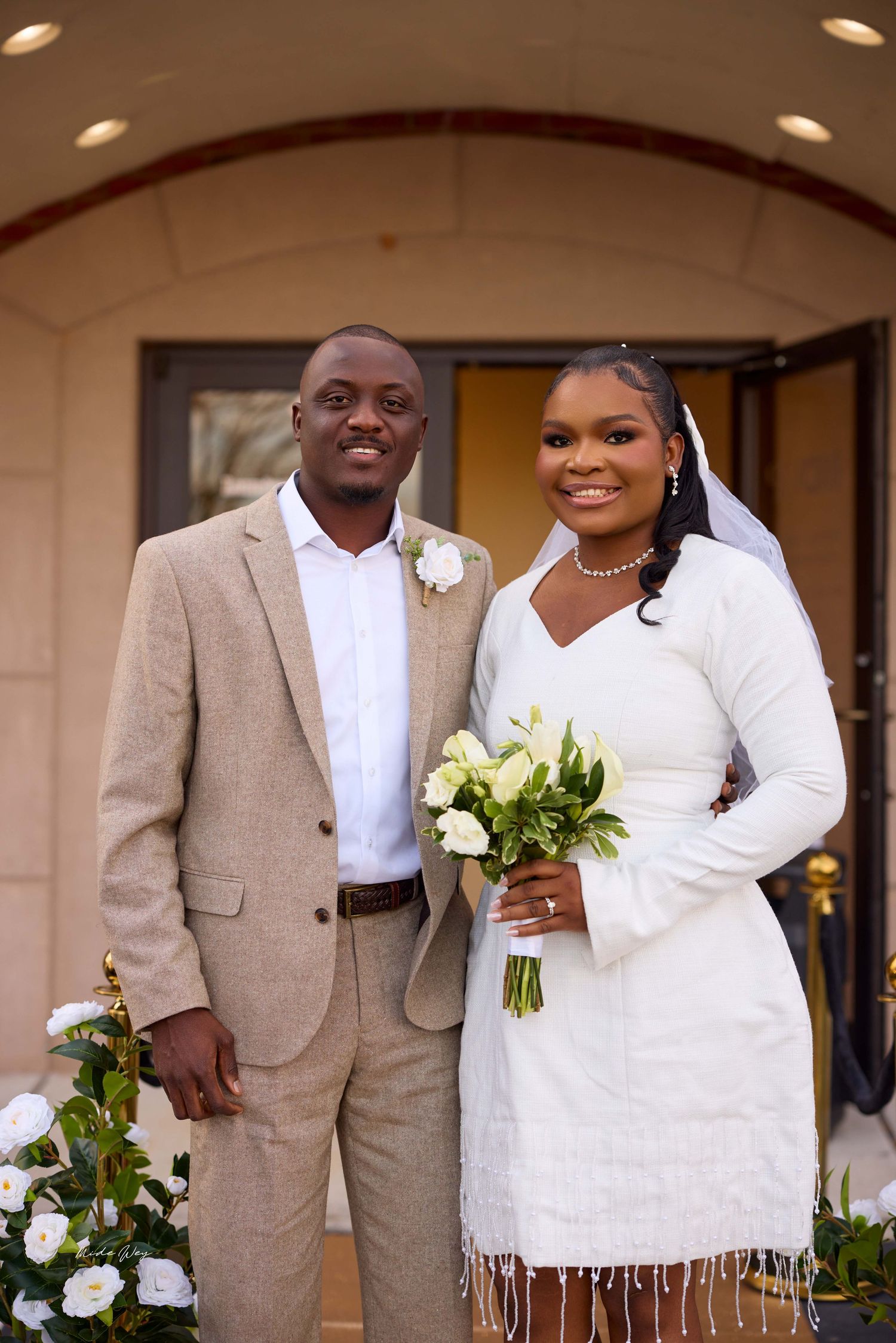 Smiling newlywed couple posing outdoors, groom in tan suit with boutonniere, bride in white dress holding bouquet.