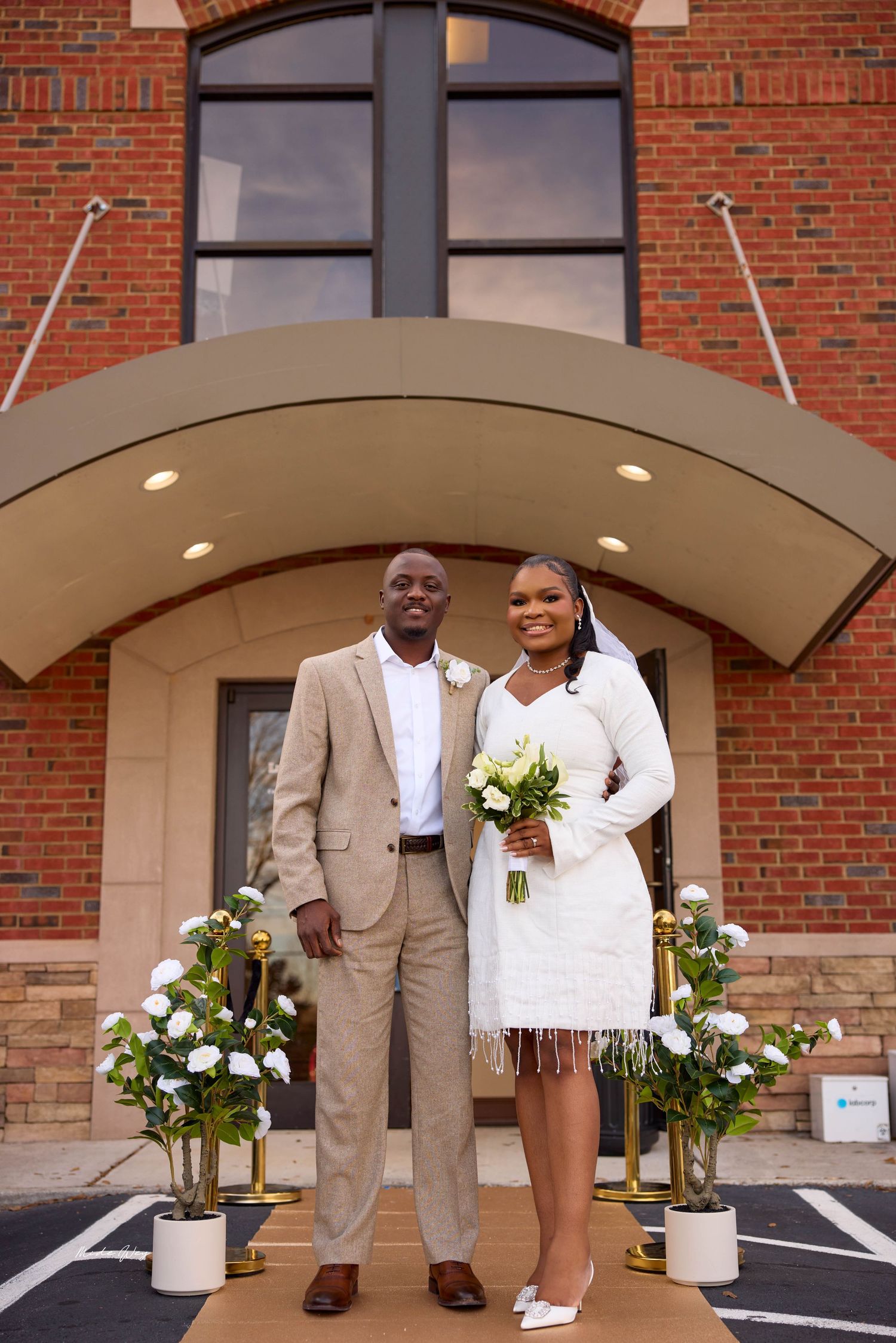 Bride and groom posing in wedding attire outside a brick building, bride holding white bouquet.