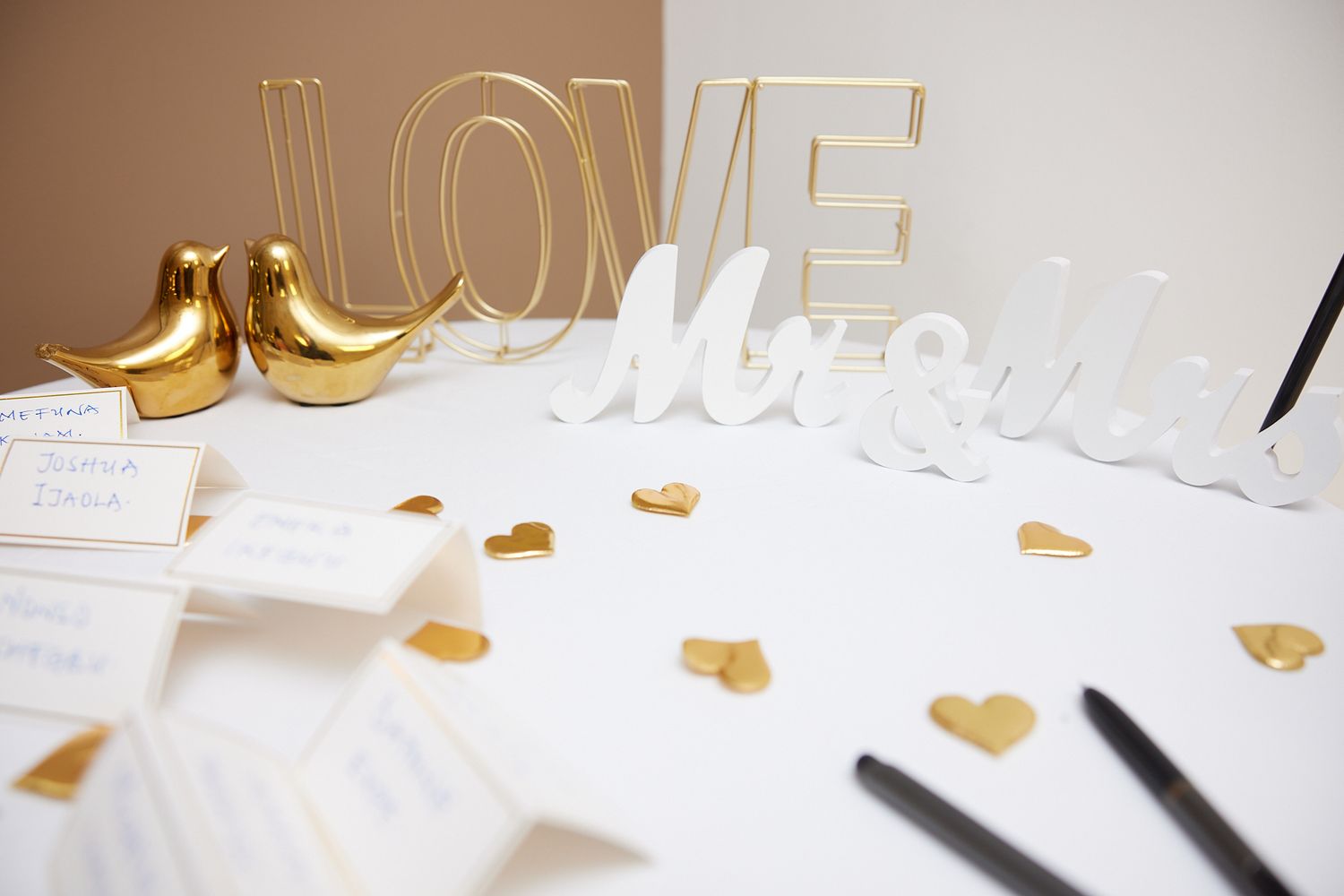 Gold LOVE letters, Mr & Mrs signs, love bird figurines, and gold heart confetti on a white wedding guest book table.