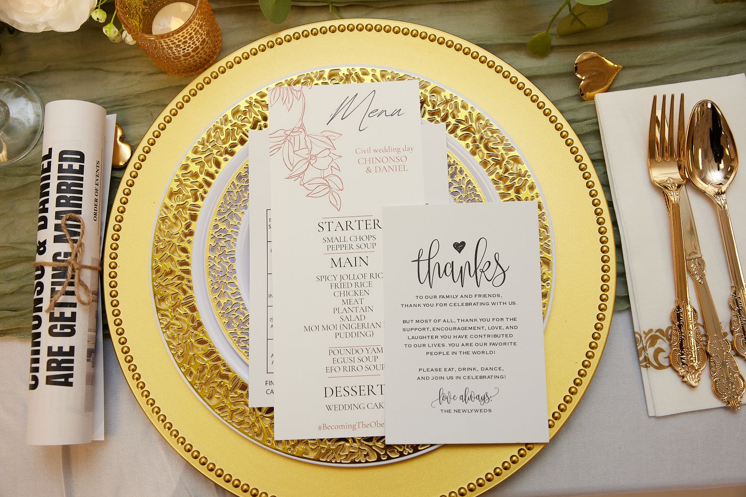 Elegant wedding place setting with gold charger plate, menu card, thank you note, and gold cutlery on white tablecloth.