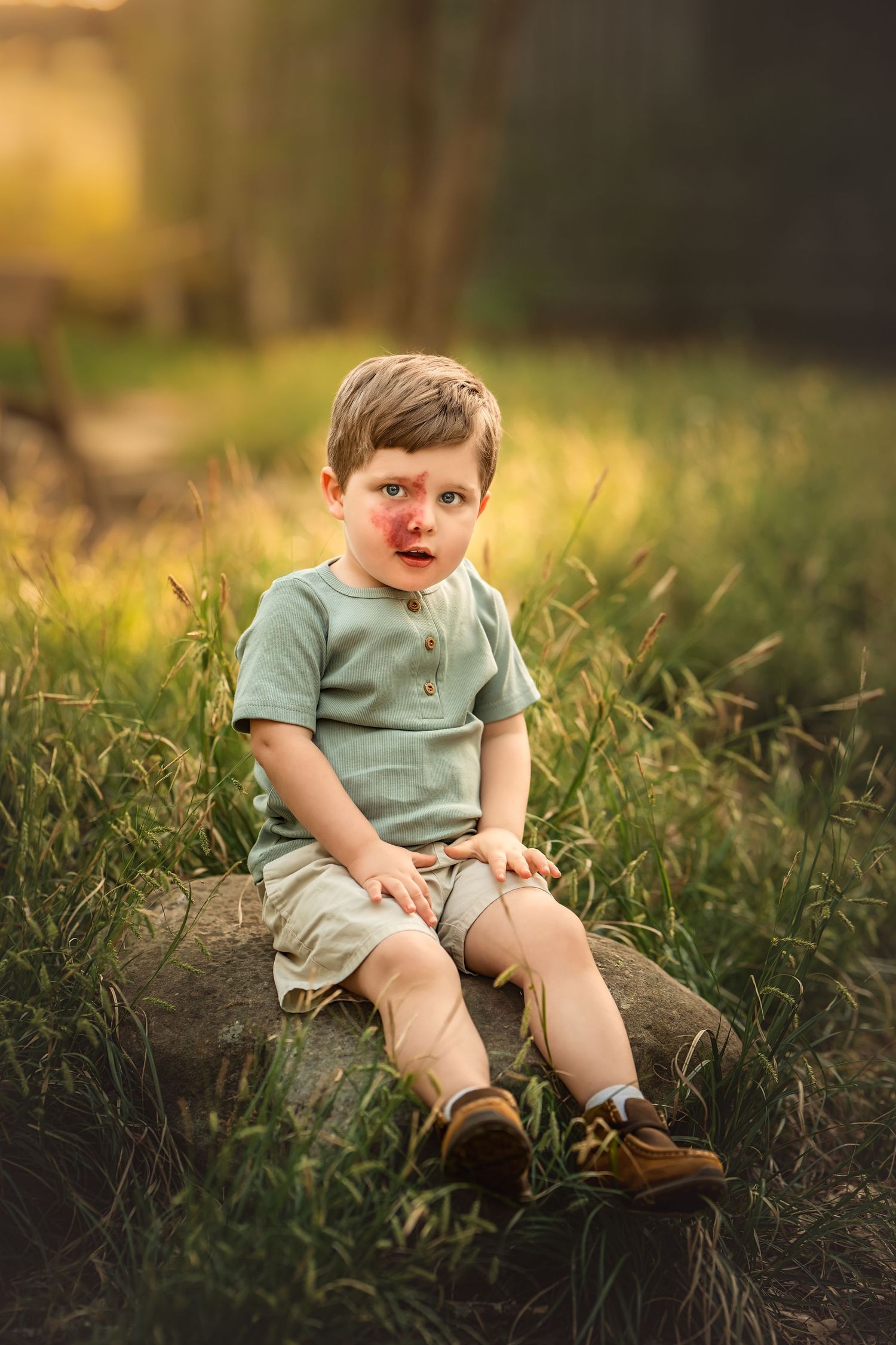 Young toddler boy sitting on a rock in a grassy field, wearing a mint green shirt and tan shorts in golden hour light.