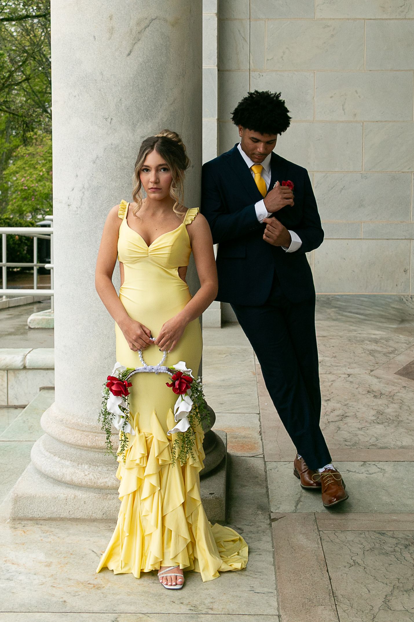 Young couple in prom attire, girl in yellow ruffled gown with floral bouquet, boy in navy suit with gold tie.