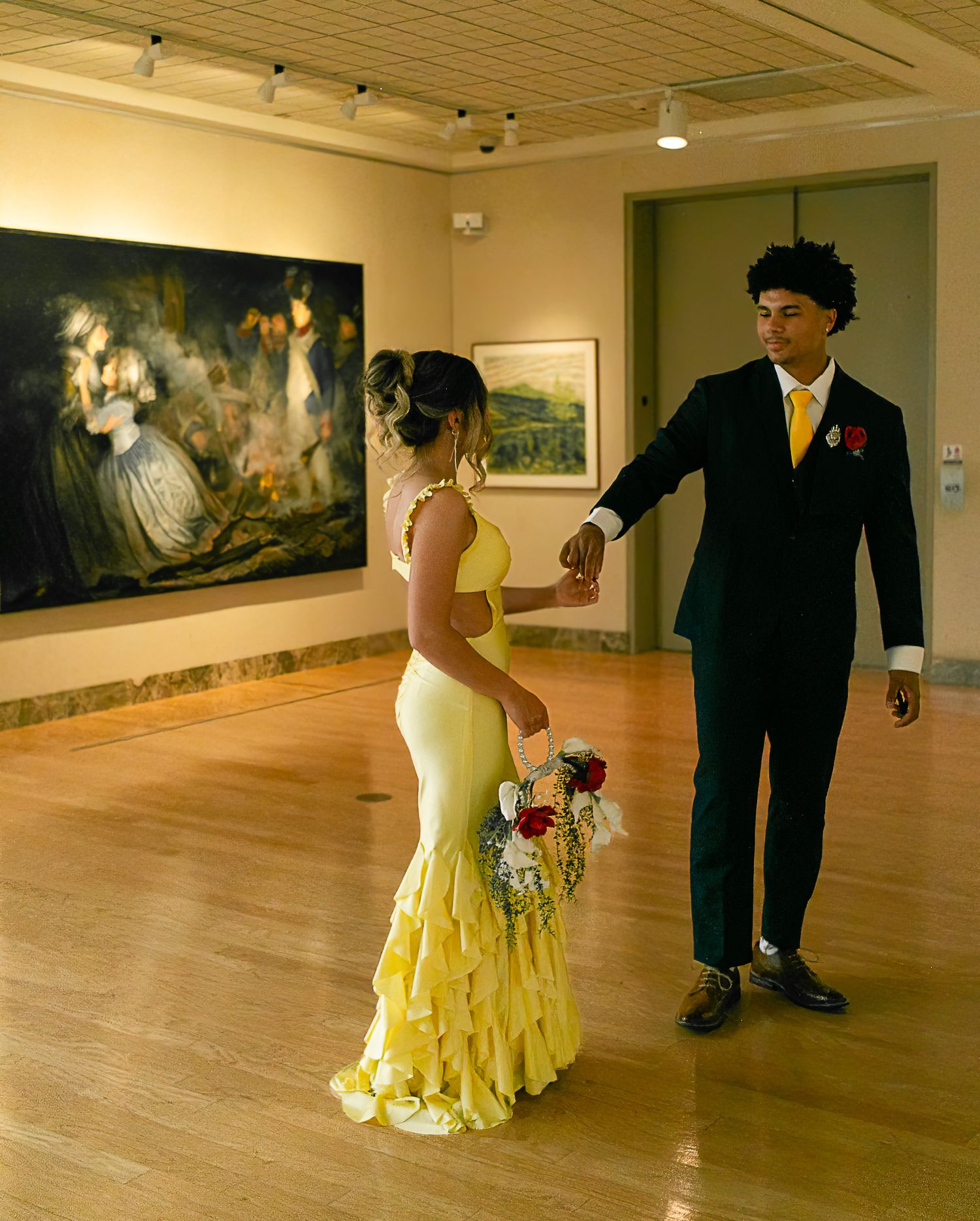 Couple in formal attire dancing in art gallery, woman in yellow ruffled gown holding bouquet, man in dark suit.