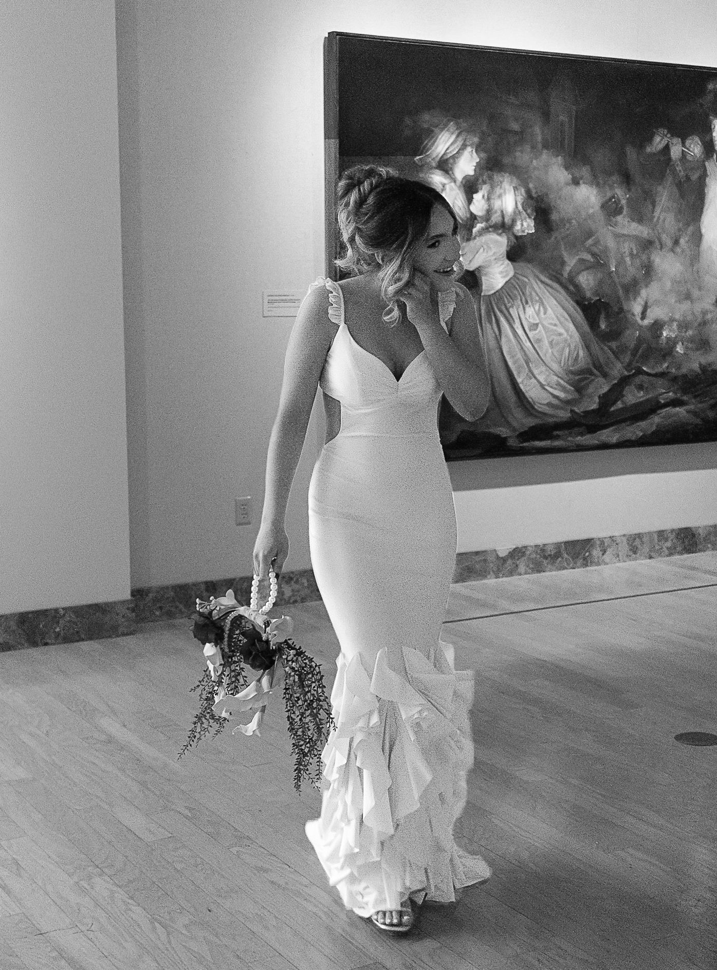 Bride in a white mermaid gown with ruffles holds a bouquet in a gallery with a classic painting backdrop.