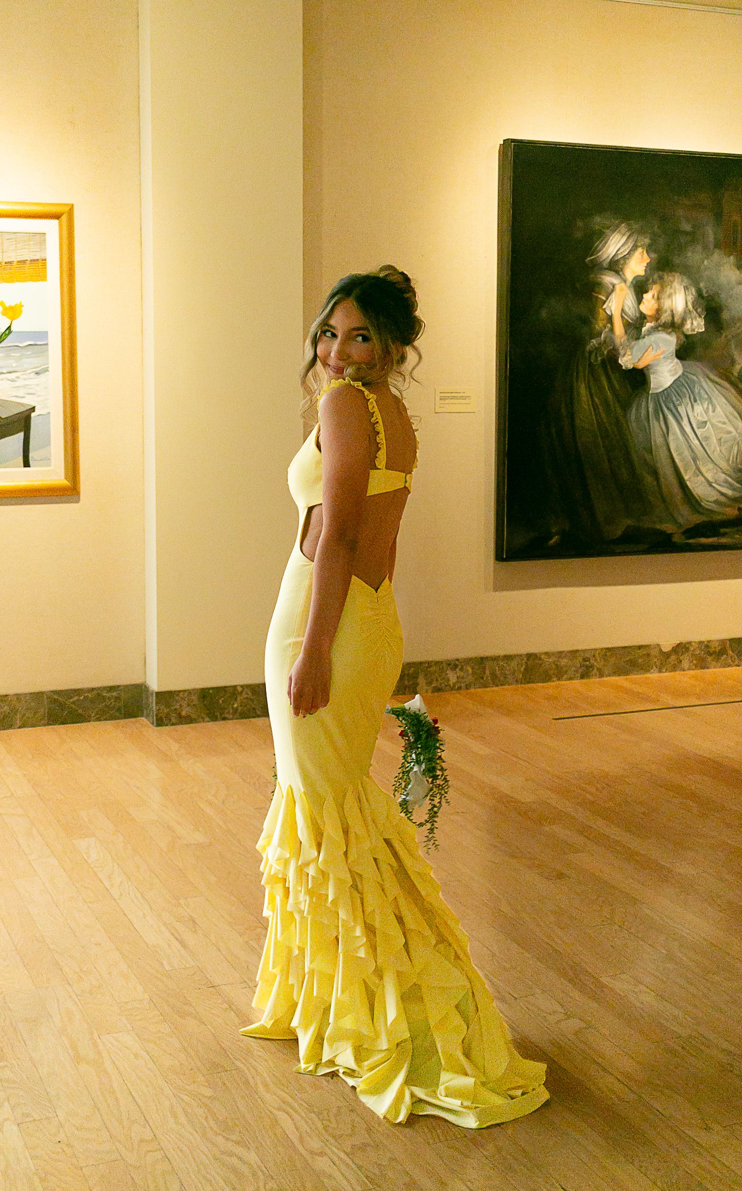 Woman in yellow mermaid gown with ruffles and open back holding bouquet in art gallery.