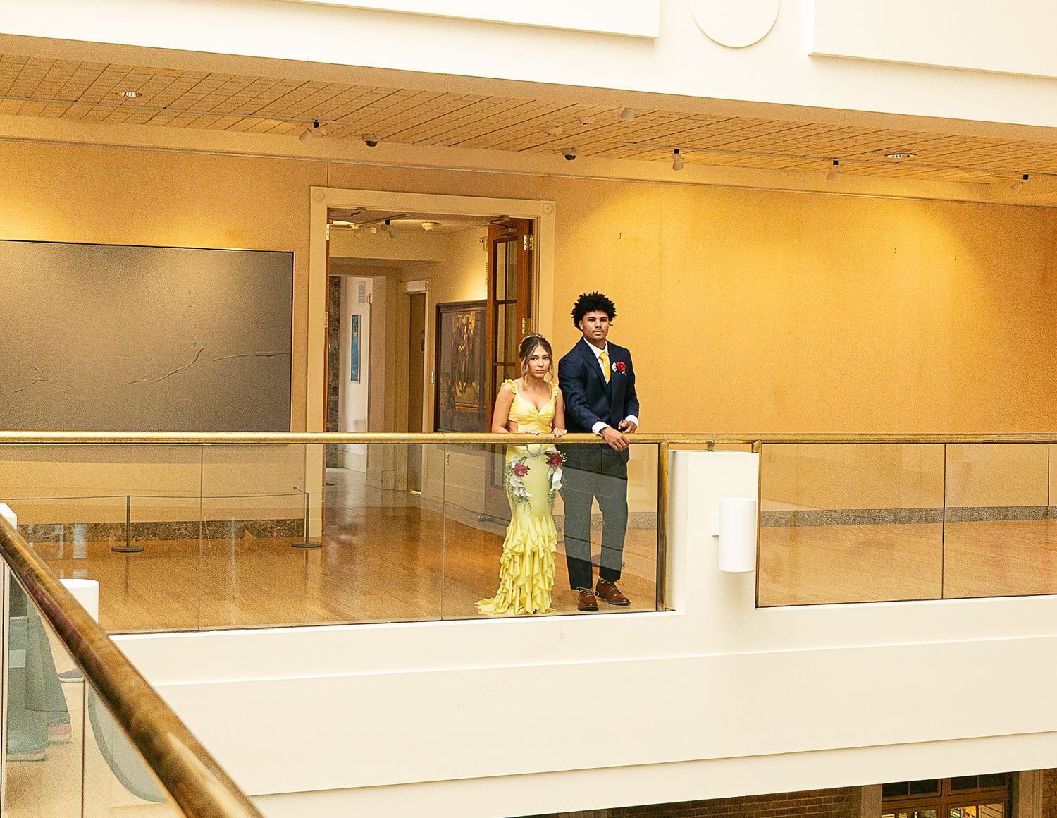 Elegant couple in formal attire standing on a balcony overlooking a modern gallery hall.