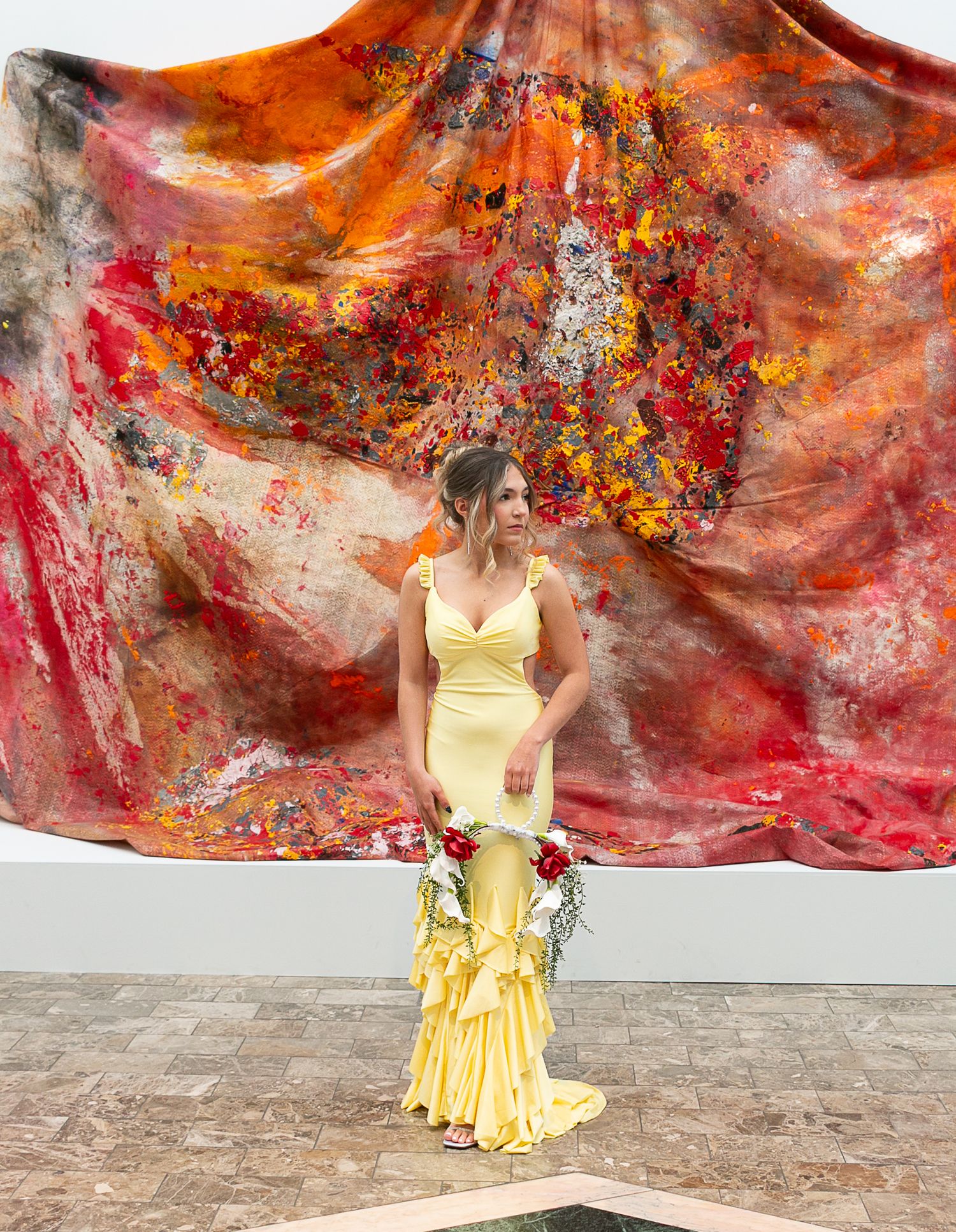 Woman in yellow ruffle gown holds red bouquet before vibrant abstract painting in red, orange, and gold.