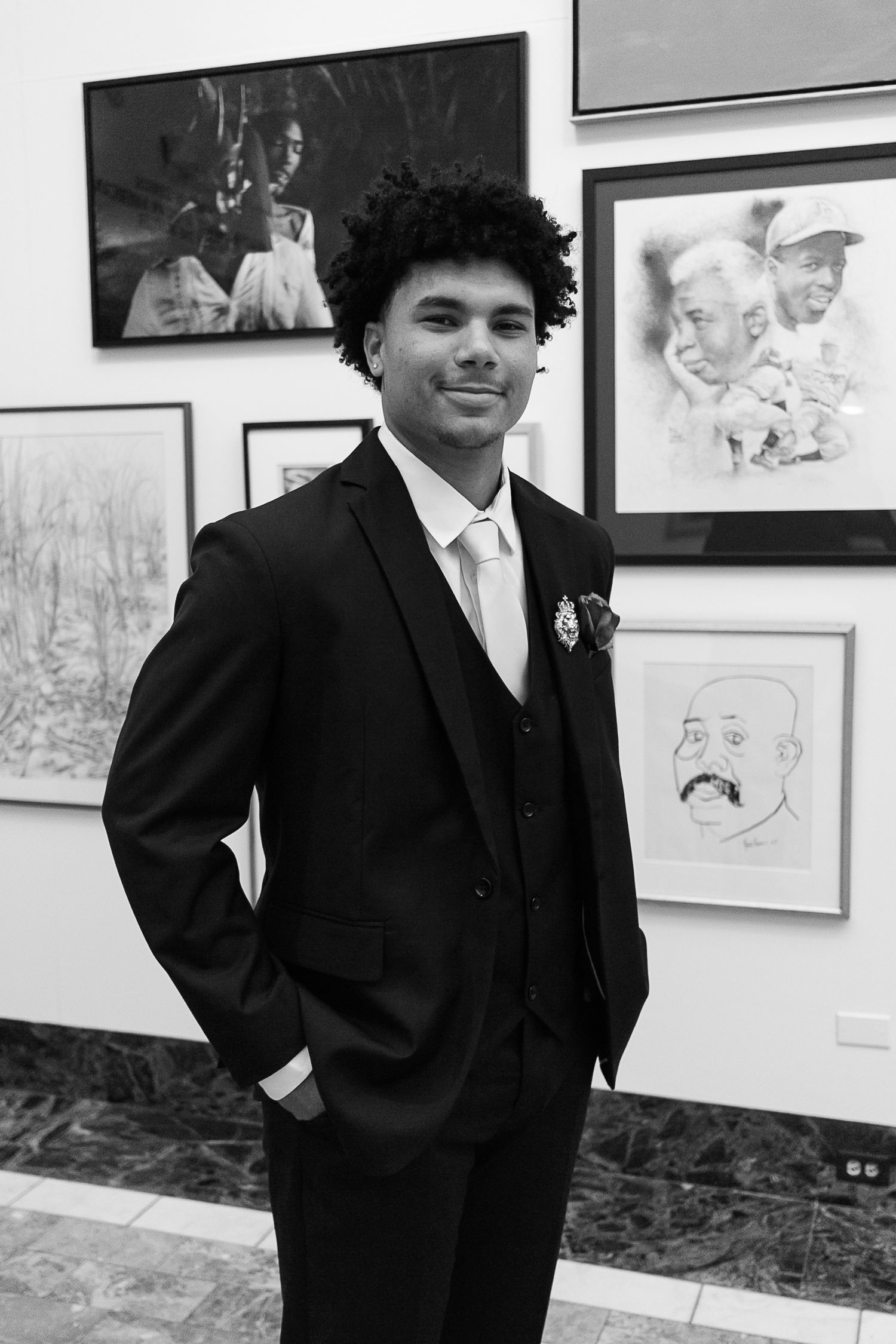 Young man in black suit with boutonniere smiling in art gallery with framed portraits on wall behind him.