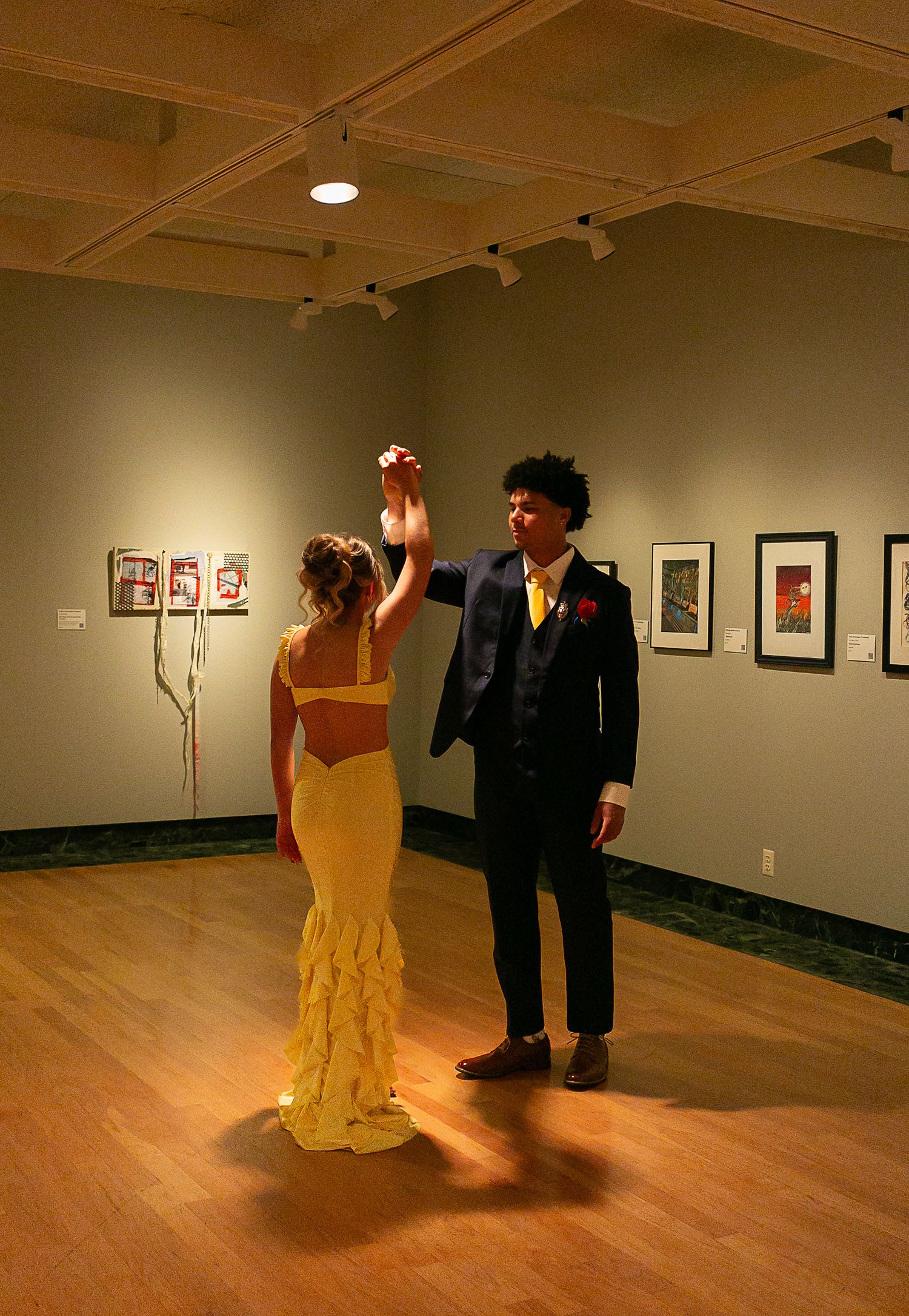 Couple dancing in art gallery, woman in yellow gown, man in navy suit, artwork displayed on walls.