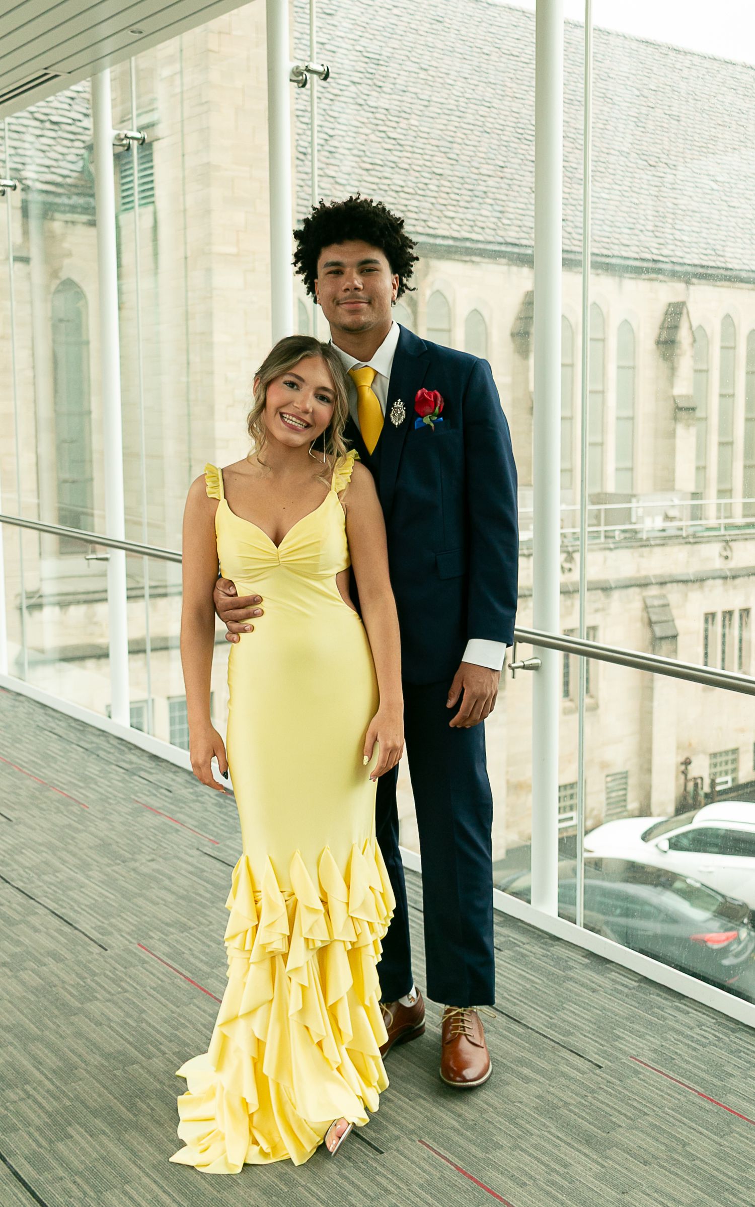 Couple posing at prom, woman in yellow mermaid gown, man in navy suit with yellow tie and red boutonniere.