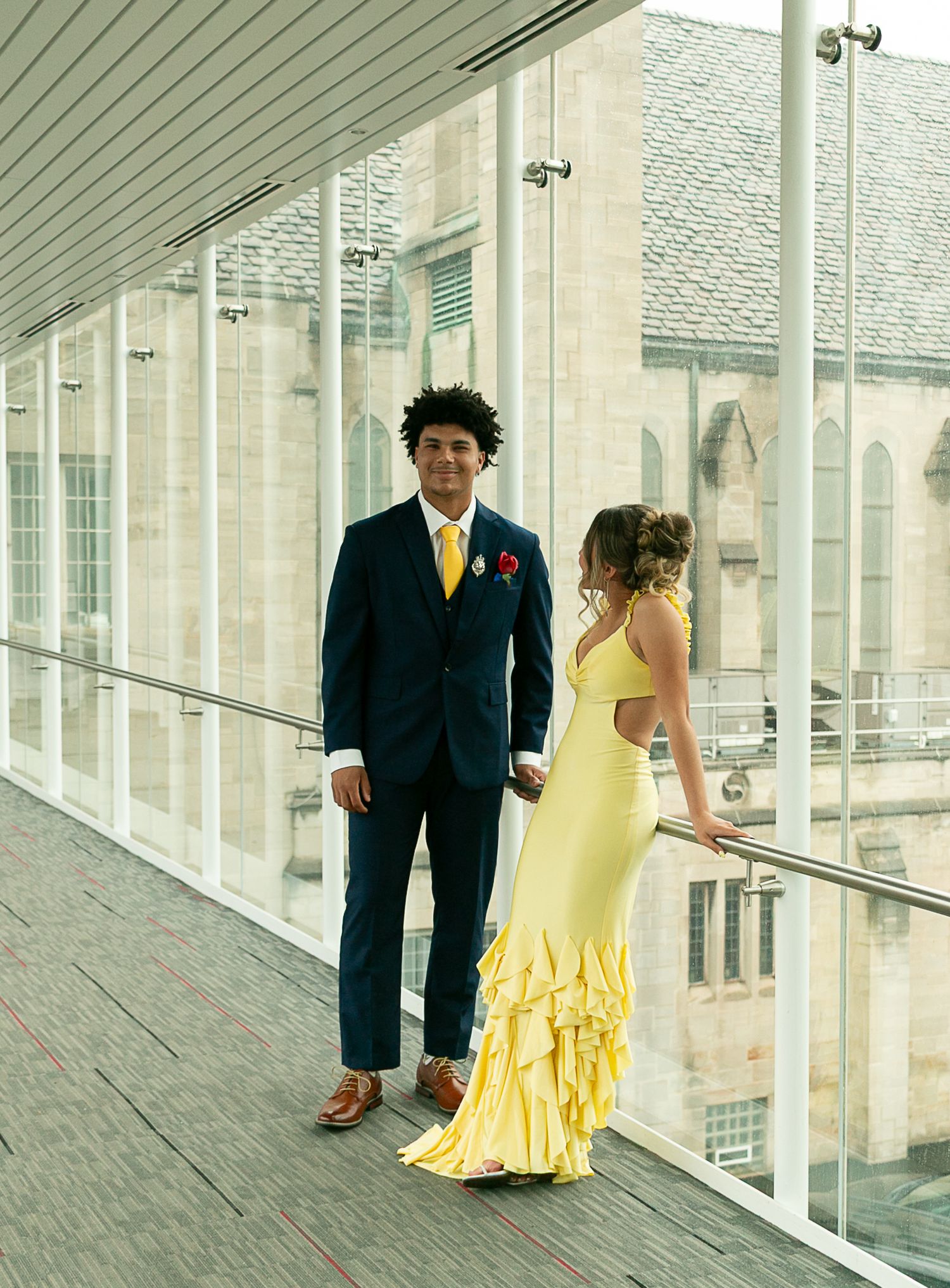 Couple in formal attire, man in navy suit with yellow tie, woman in yellow ruffled gown, posing on modern walkway.