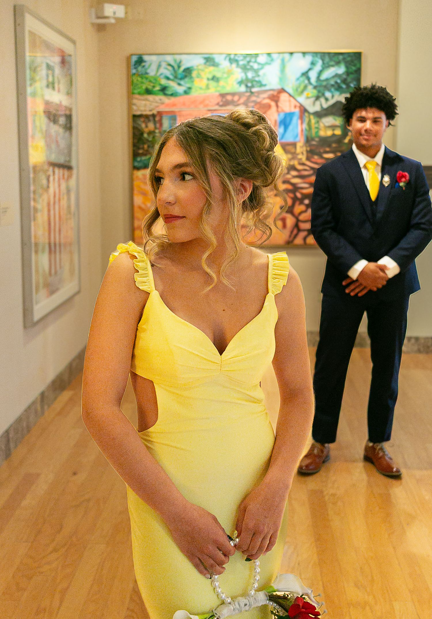 Young woman in yellow dress and man in navy suit pose in art gallery for prom photos.
